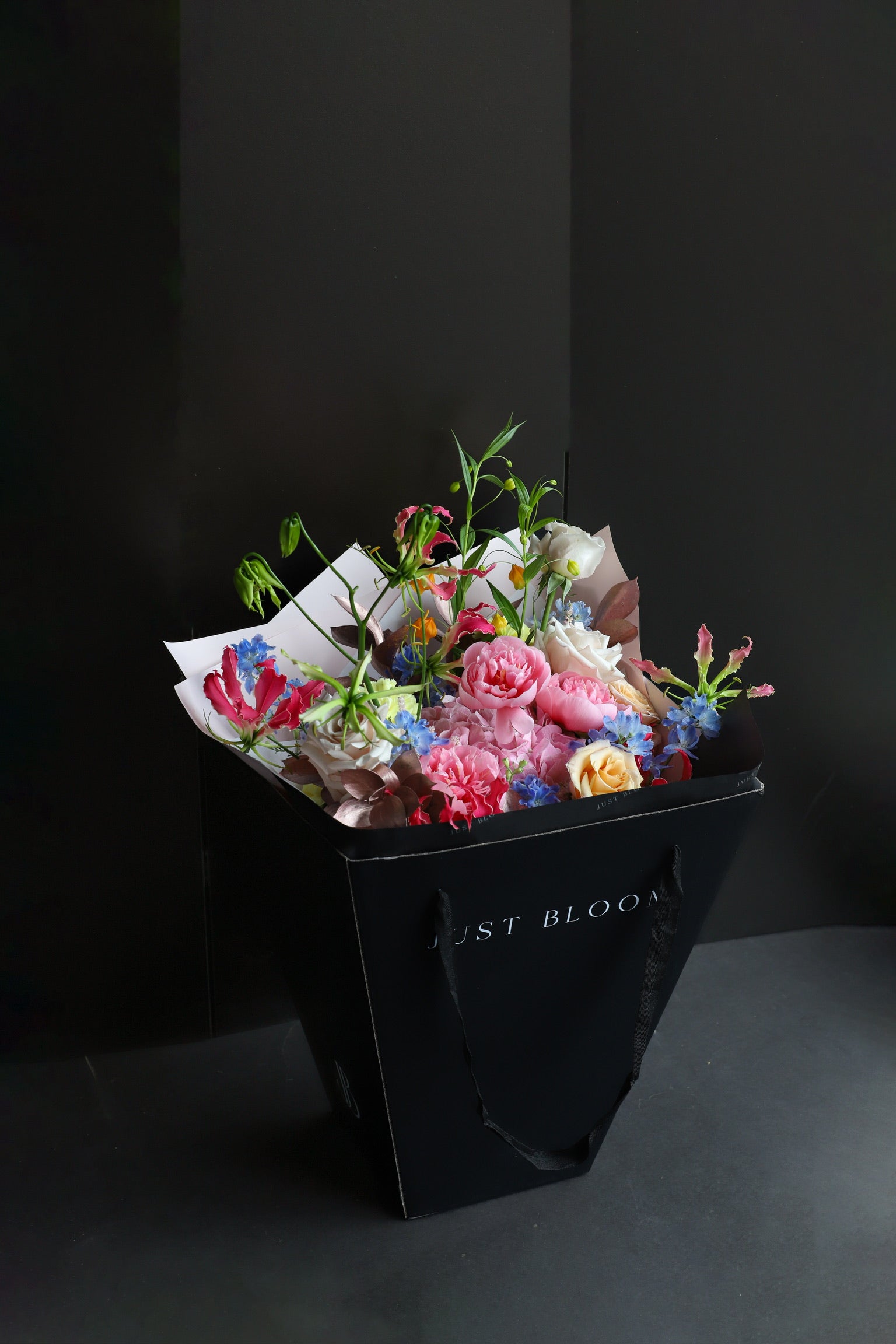 The Wild Bunch luxury bouquet by Just Bloom Hong Kong presented in a black Just Bloom tote bag, showcasing vibrant mixed blooms in pink, blue, white and orange tones