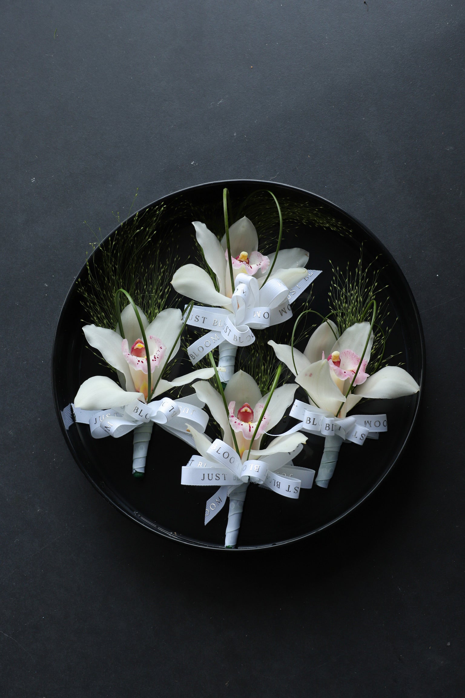 Four Boutonnieres arranged on a black tray featuring white cymbidium orchids, frosted explosion grass and steel grass tied with Just Bloom white ribbon — Just Bloom Hong Kong
