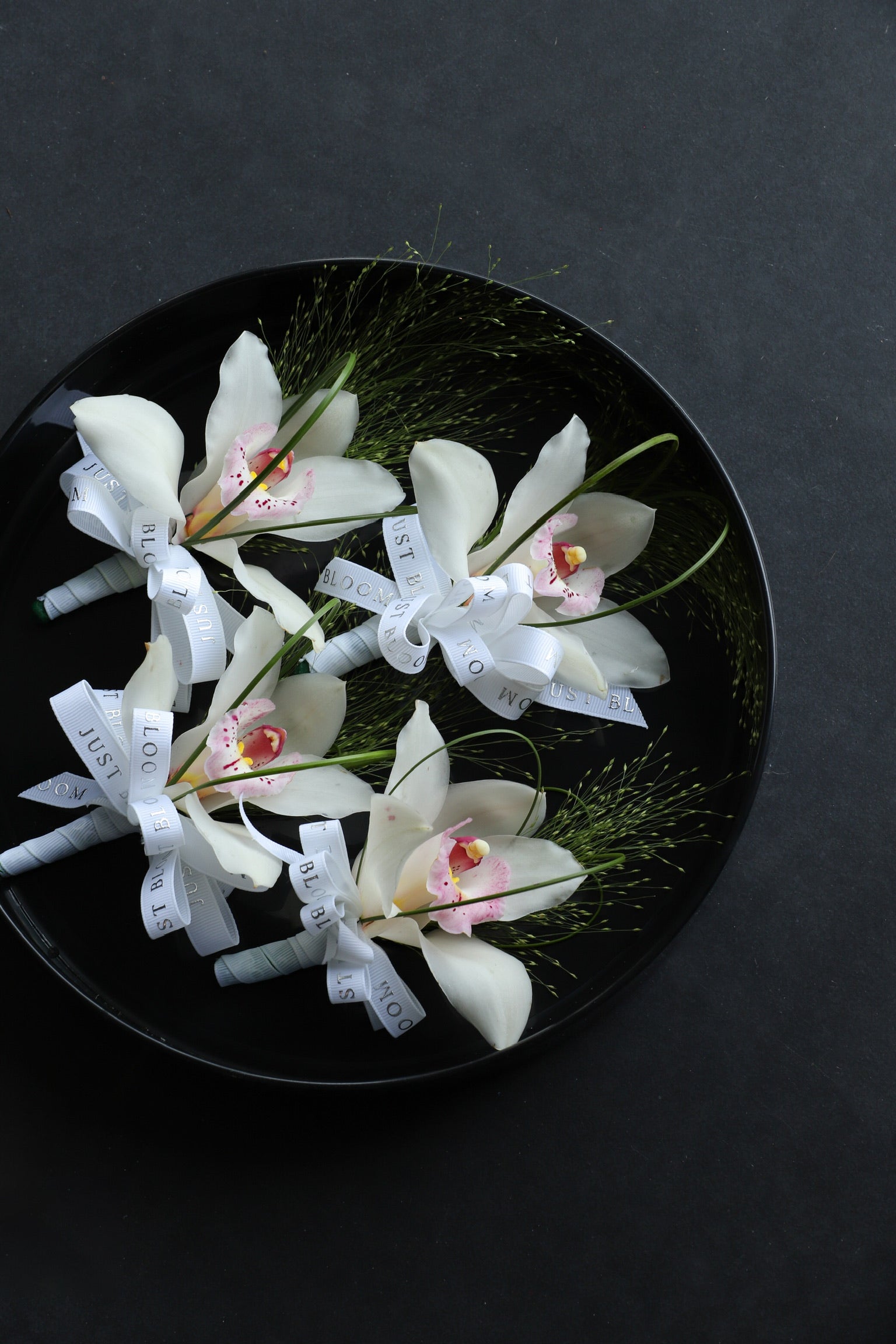 Close-up of four With Grace wedding corsages on a black tray by Just Bloom Hong Kong, showcasing white cymbidium orchids with frosted explosion grass, steel grass and Just Bloom white ribbon