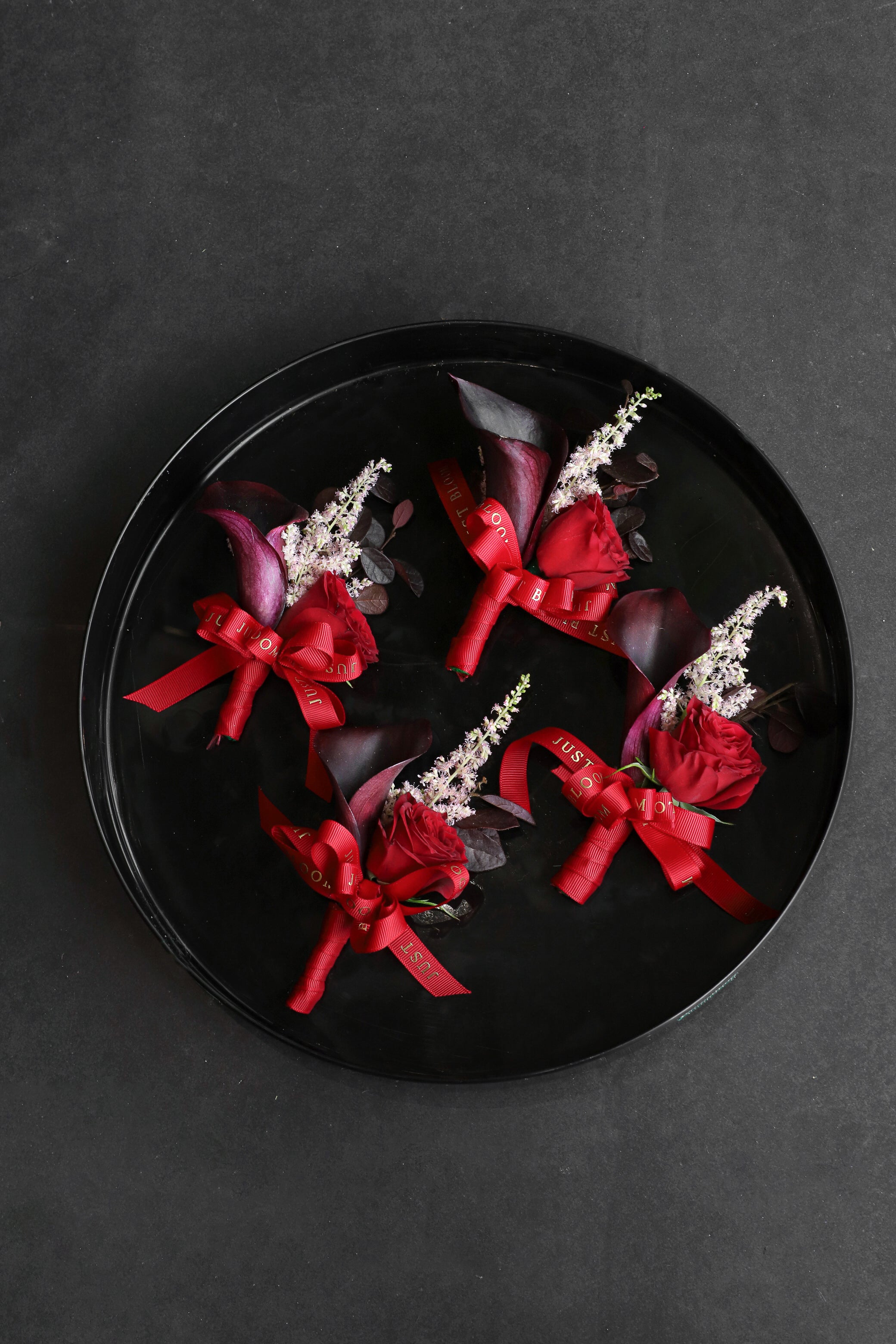 Four Boutonnieres displayed on a black tray by Just Bloom Hong Kong, showcasing deep red spray roses, dark calla lilies, astilbes and Just Bloom red ribbon