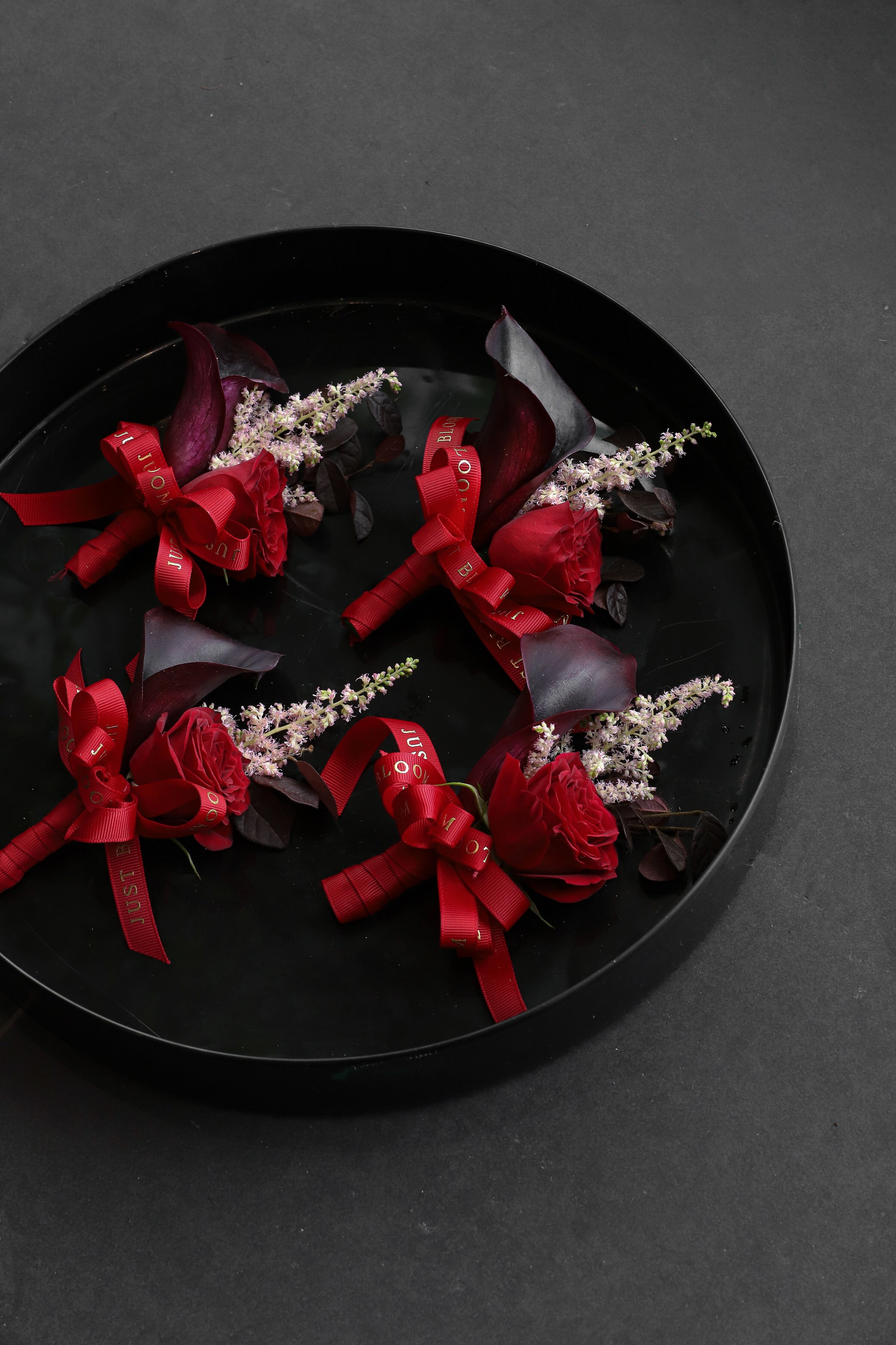 Four Boutonnieres arranged on a black tray featuring deep red spray roses, dark calla lilies and astilbes tied with Just Bloom red ribbon — Just Bloom Hong Kong