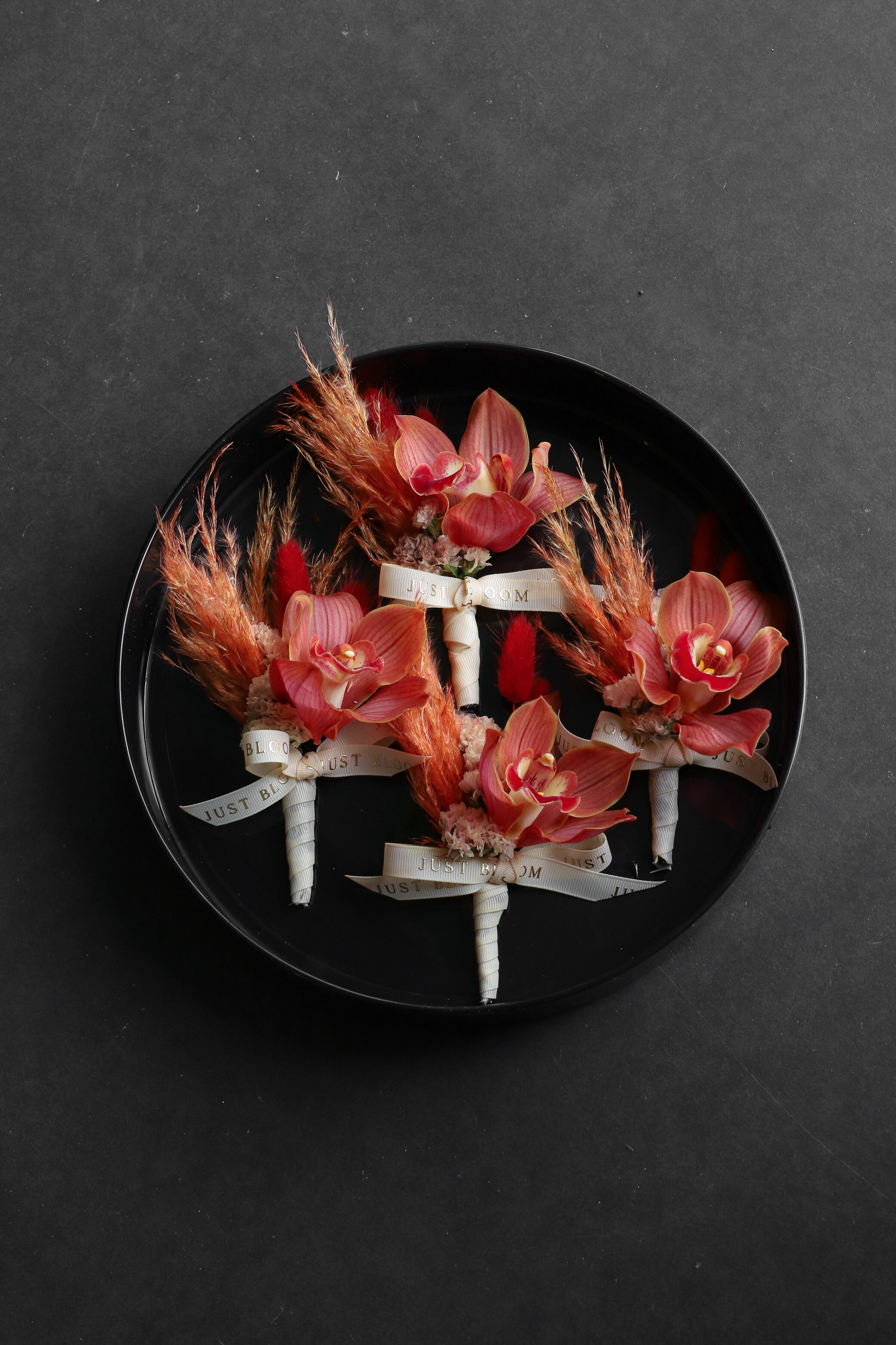 Close-up of four Boutonnieres on a black tray by Just Bloom Hong Kong, showcasing cymbidium orchids with myosotis, dried lagurus ovatus and Just Bloom ivory ribbon