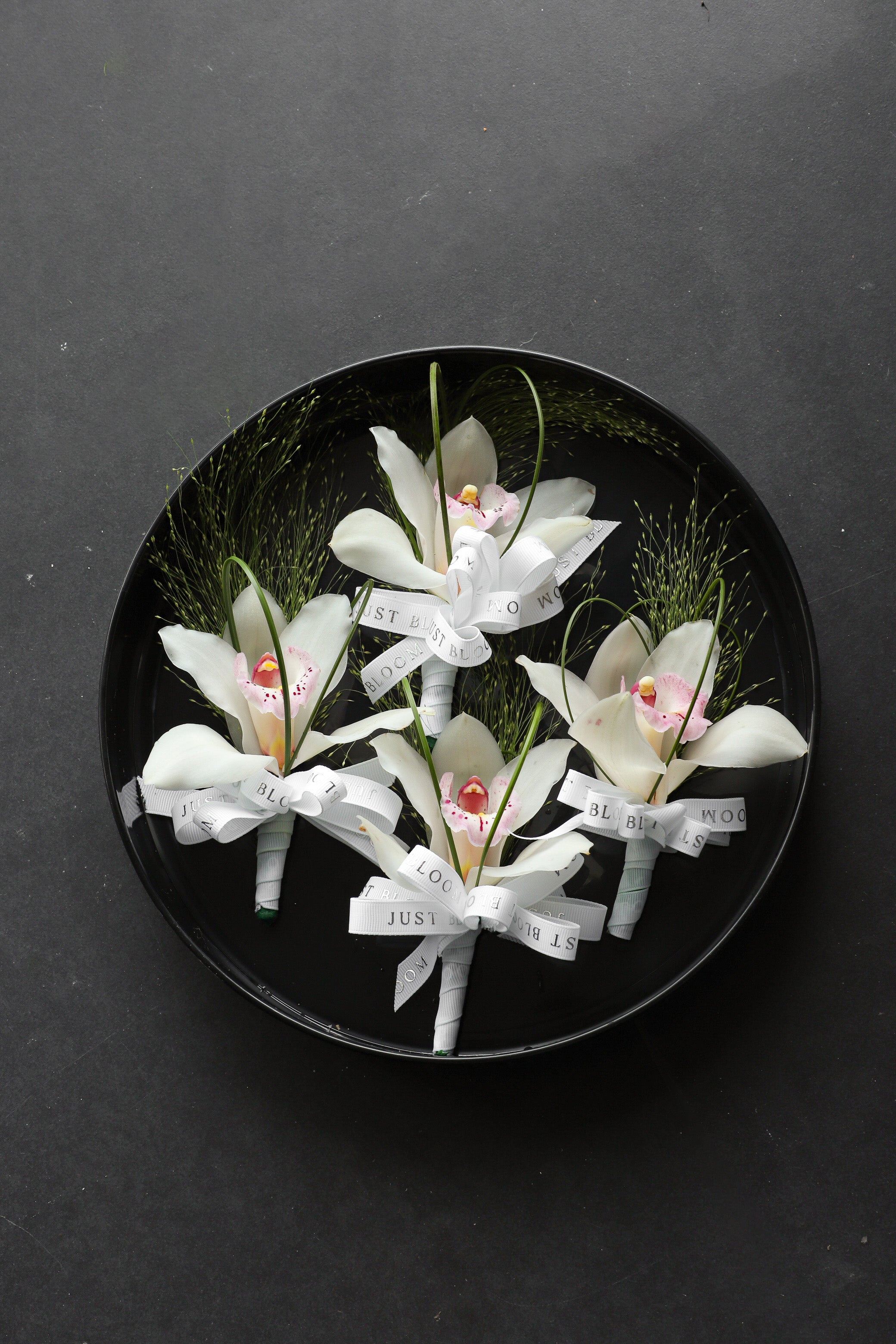 Four Boutonnieres arranged on a black tray featuring white cymbidium orchids, frosted explosion grass and steel grass tied with Just Bloom white ribbon — Just Bloom Hong Kong
