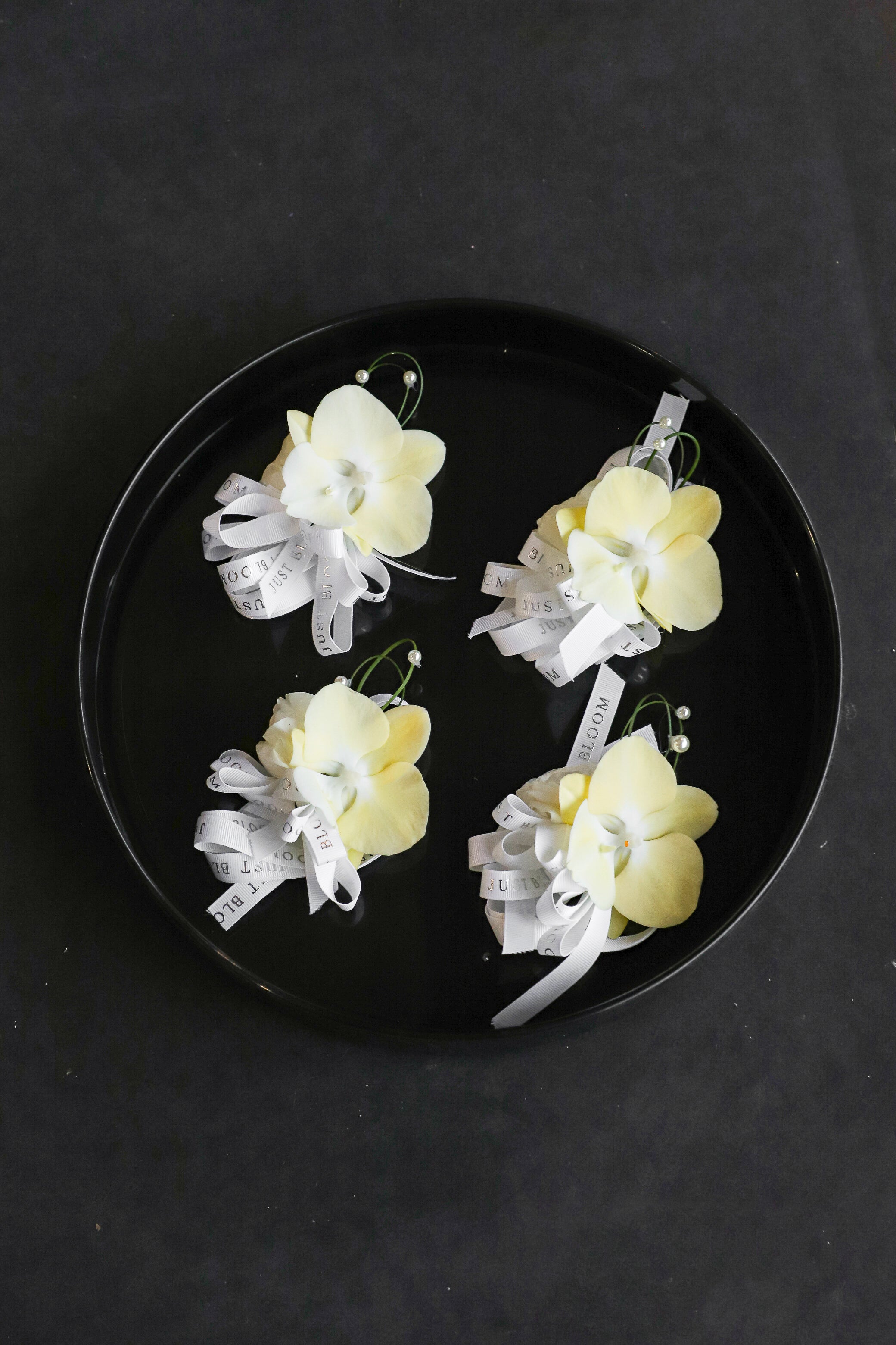 Four Meant to Be corsages arranged on a black tray featuring cream phalaenopsis orchids, steel grass and pearl accents tied with Just Bloom white ribbon — Just Bloom Hong Kong