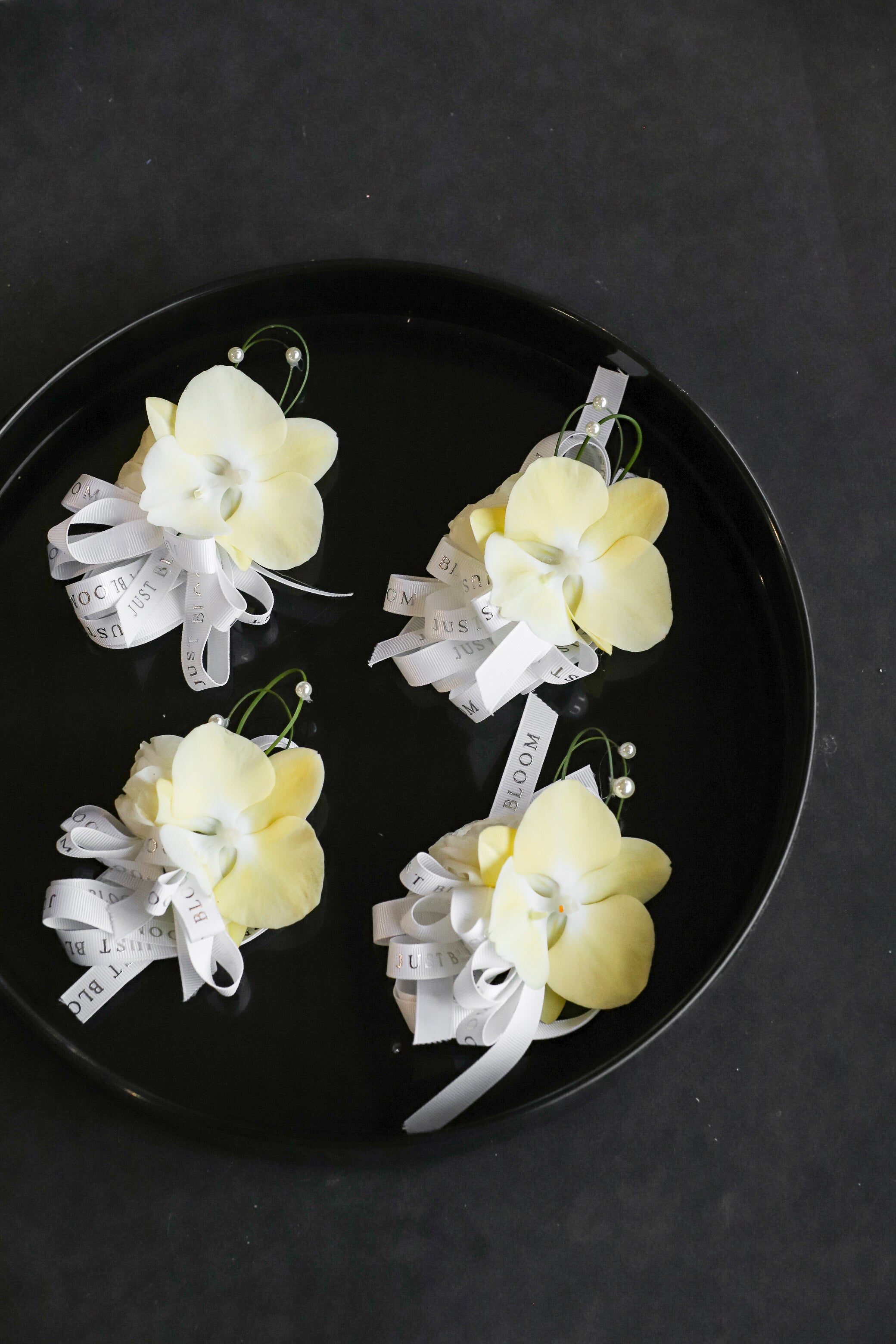 Close-up of four Meant to Be wedding corsages on a black tray by Just Bloom Hong Kong, showcasing cream phalaenopsis orchids with spiral steel grass, pearl accents and Just Bloom white ribbon