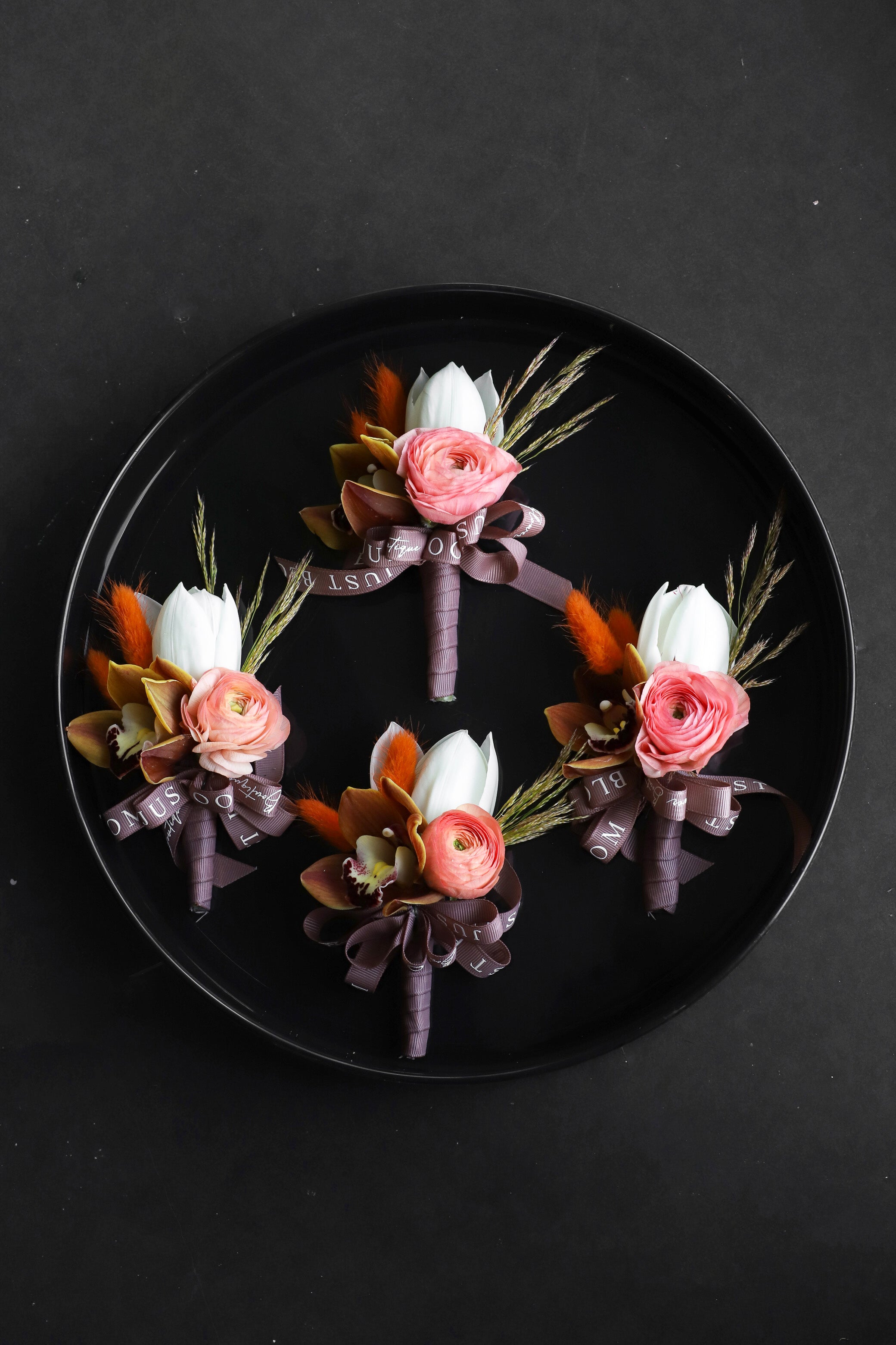 Close-up of four Meant to Be wedding corsages on a black tray by Just Bloom Hong Kong, showcasing cream phalaenopsis orchids with spiral steel grass, pearl accents and Just Bloom white ribbon