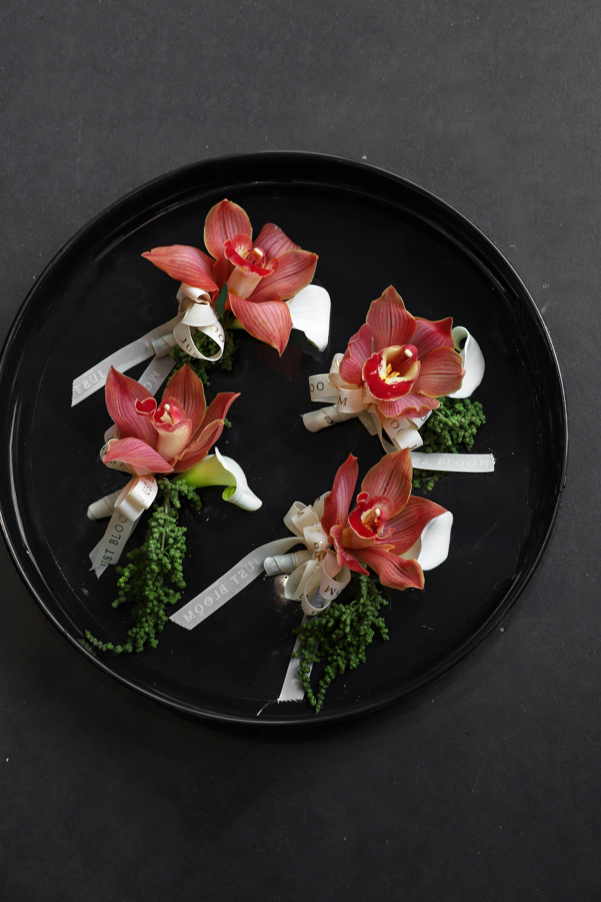Four Amber Boutonniere arranged on a black tray featuring terracotta cymbidium orchids, white calla lilies and foliage tied with Just Bloom ivory ribbon — Just Bloom Hong Kong