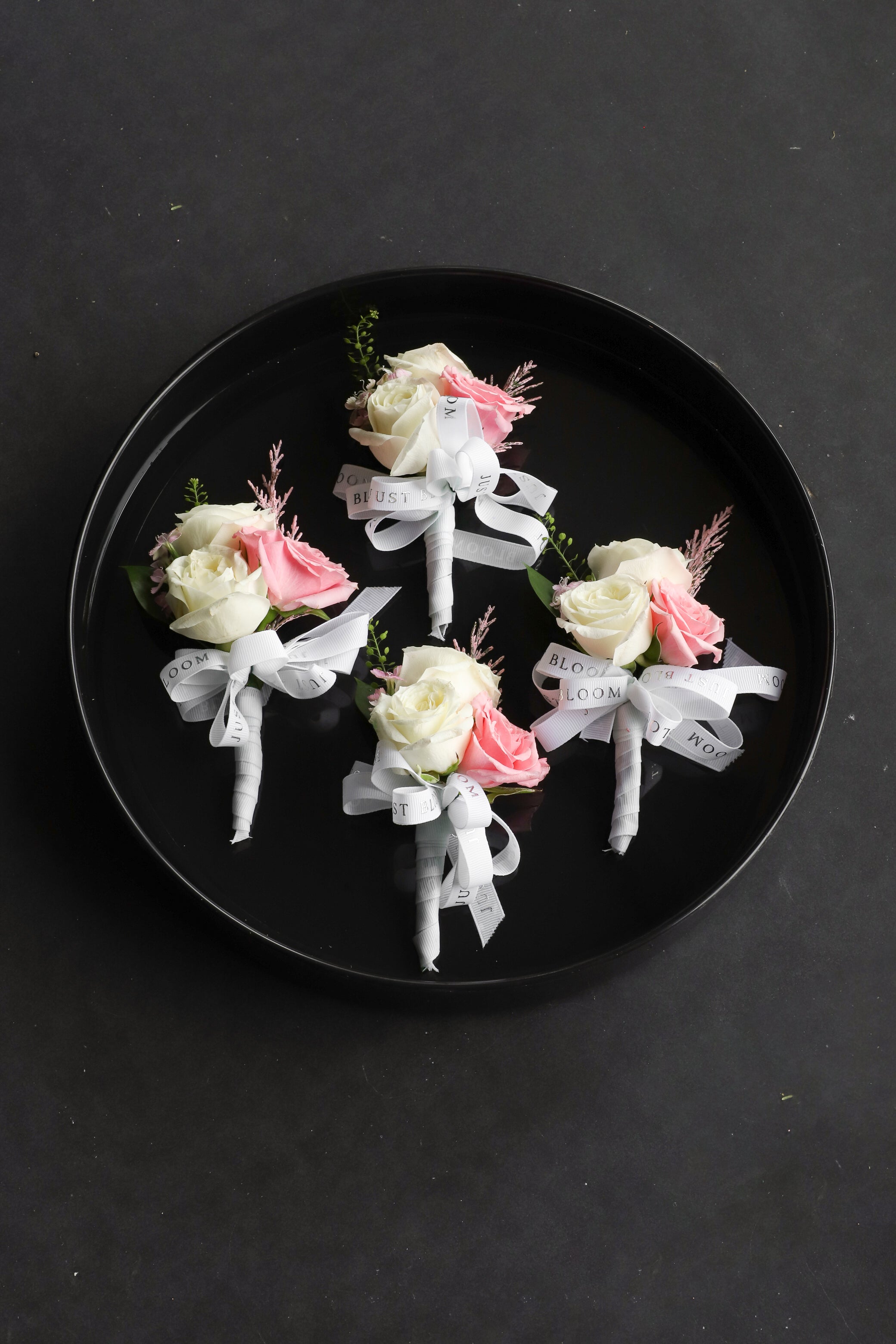 Four Boutonnieres arranged on a black tray featuring white and pink Ecuadorian roses, frosted explosion and astilbes tied with Just Bloom white ribbon — Just Bloom Hong Kong