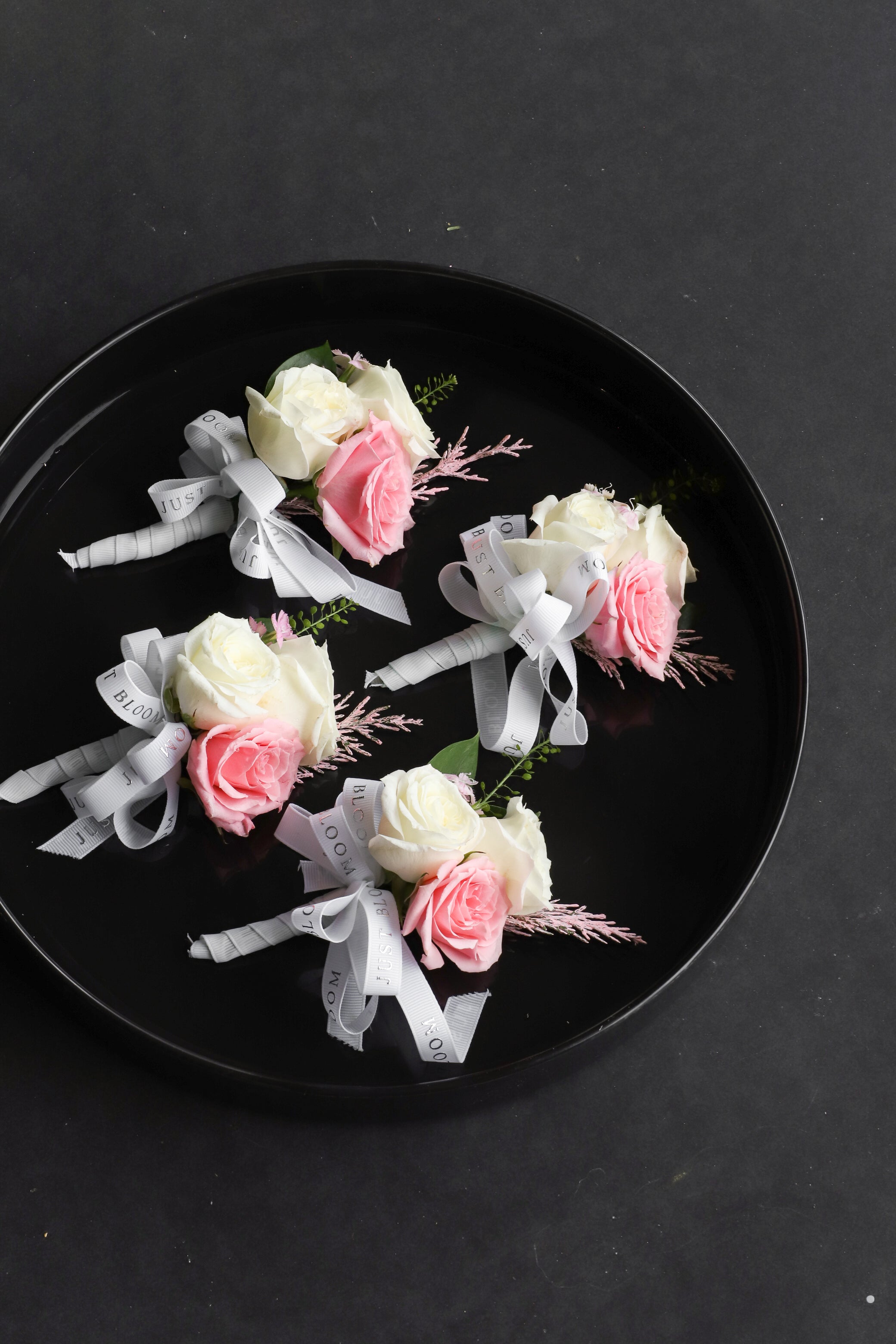 Close-up of Boutonnieres on a black tray by Just Bloom Hong Kong, showcasing white and pink Ecuadorian roses with frosted explosion, astilbes and Just Bloom white ribbon