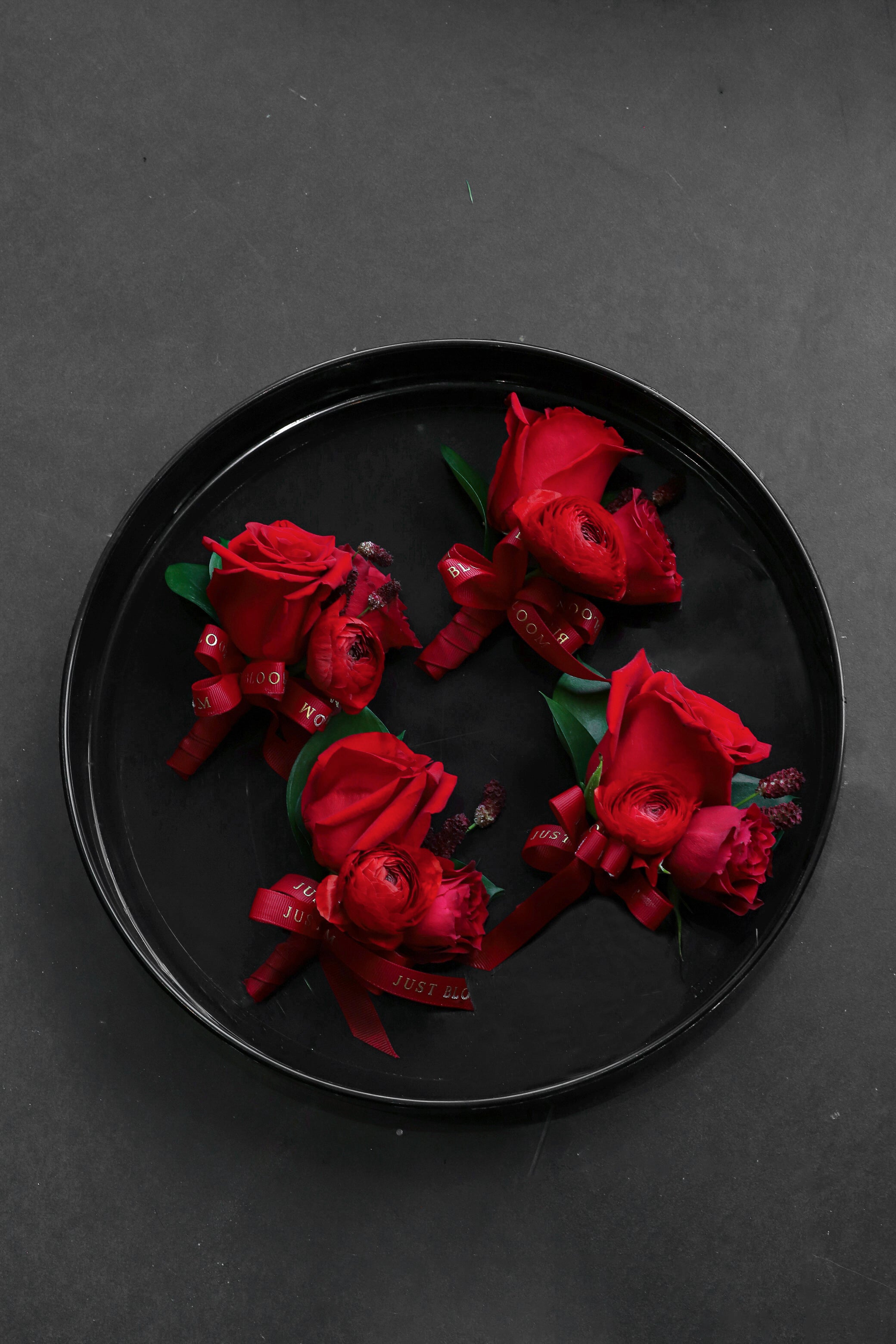 Four True Red Boutonnieres arranged on a black tray featuring deep red Ecuadorian roses, spray roses, ranunculus and seasonal berries tied with Just Bloom deep red ribbon — Just Bloom Hong Kong