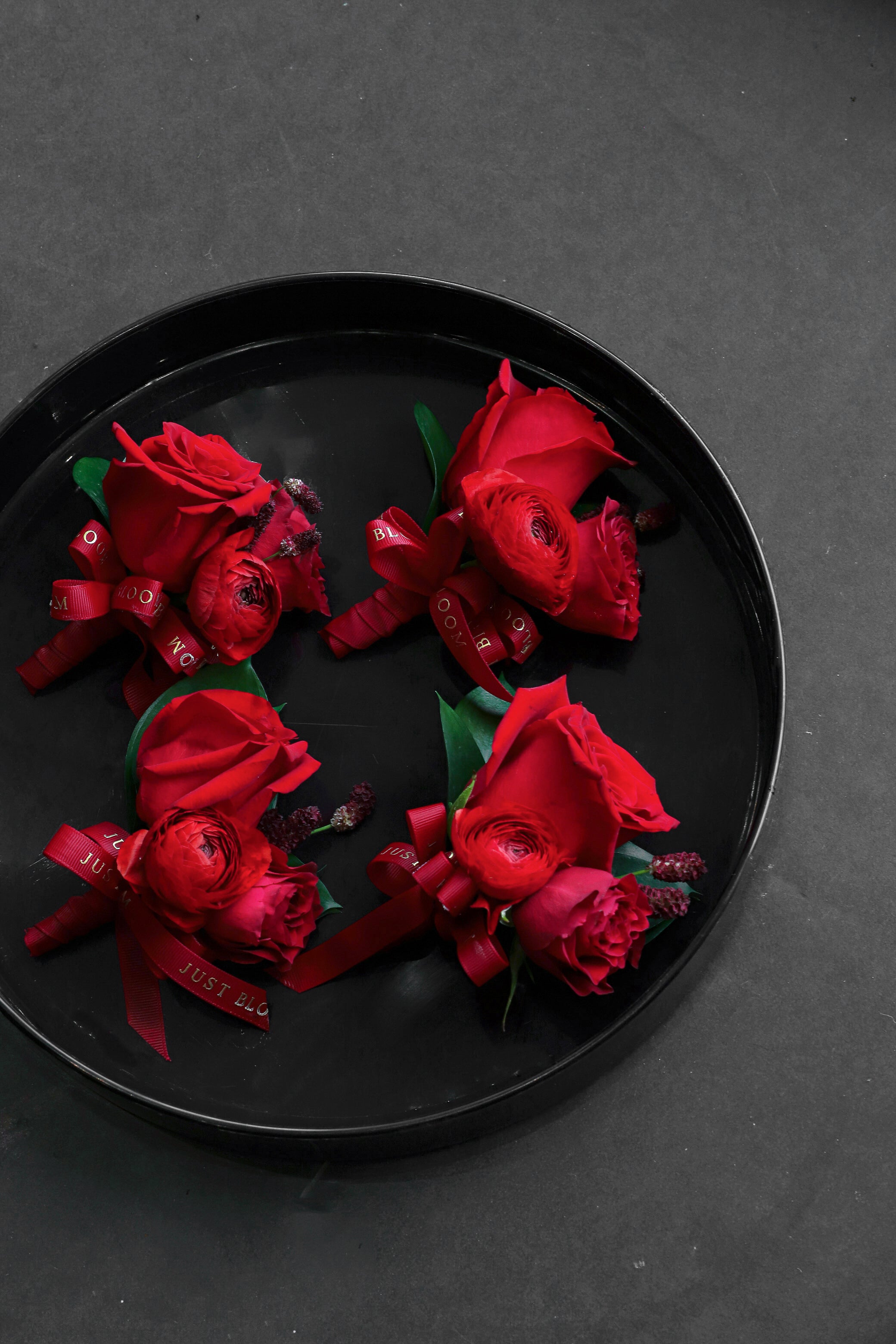 Close-up of four True Red wedding corsages on a black tray by Just Bloom Hong Kong, showcasing deep red roses, ranunculus and seasonal berries with Just Bloom deep red ribbon