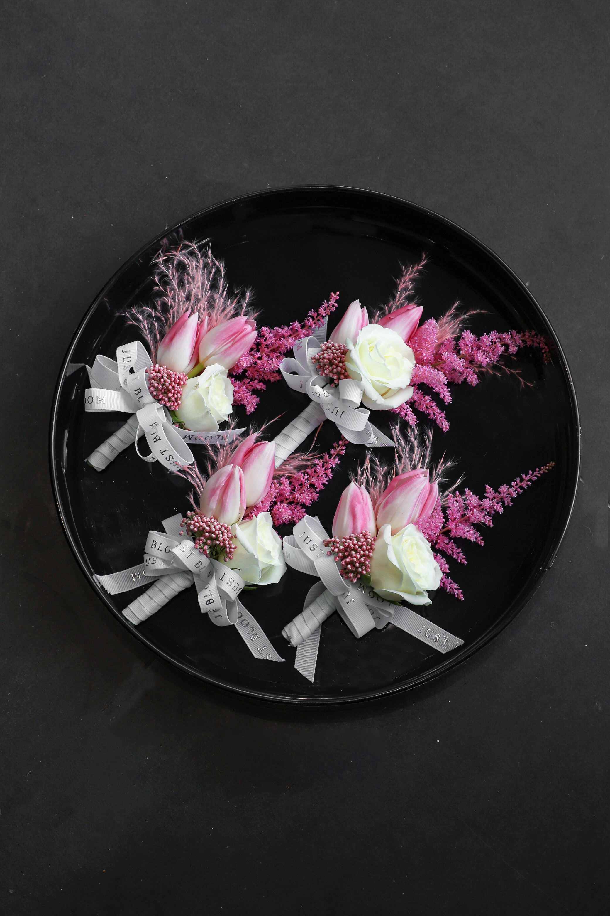 Close-up of four Sweet Vow Boutonnieres on a black tray by Just Bloom Hong Kong, showcasing pink tulips, white spray roses, deep pink astilbes and delicate rice flowers with Just Bloom grey ribbon