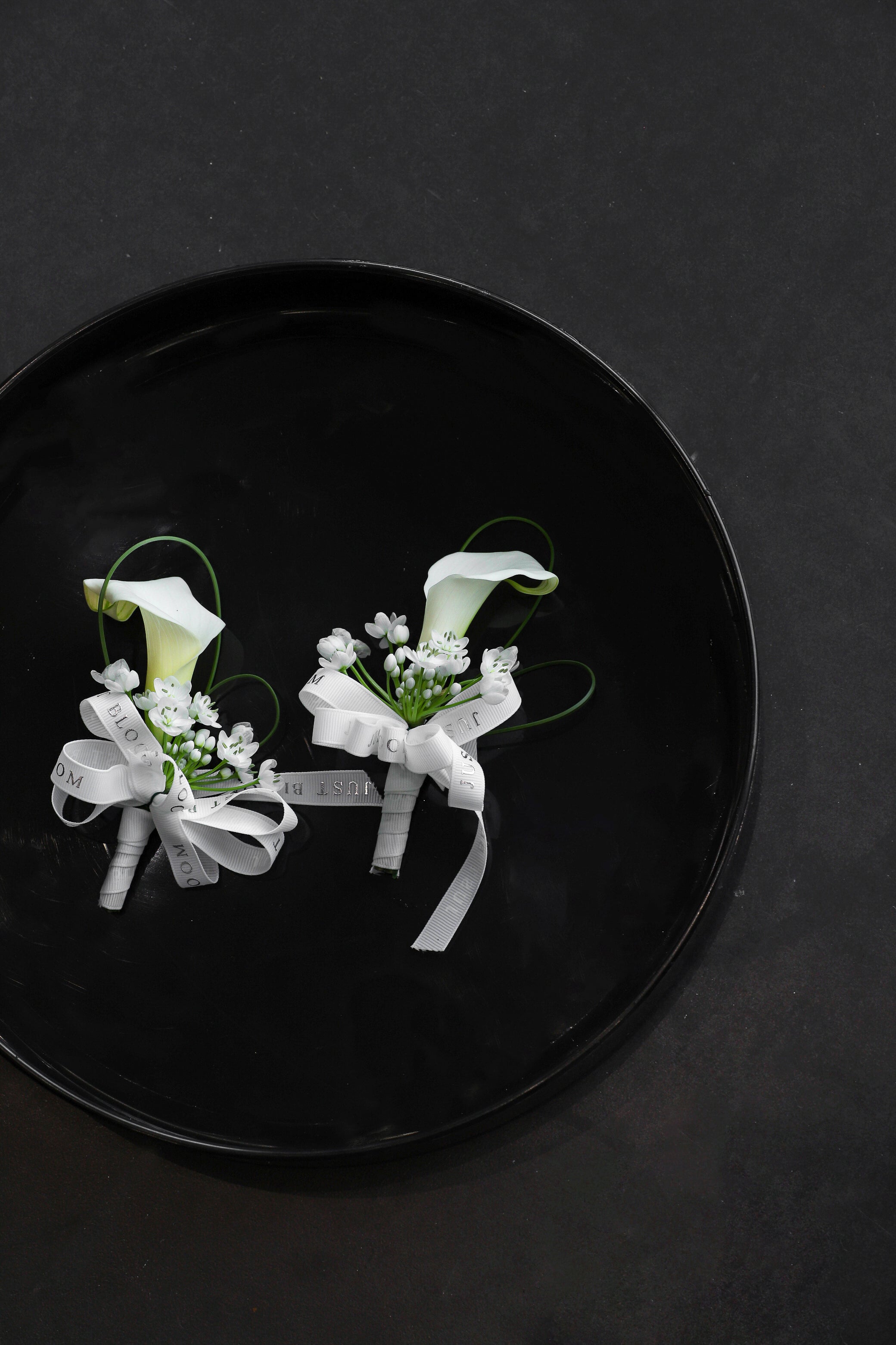 Close-up of two Simply White wedding Boutonnieres on a black tray by Just Bloom Hong Kong, showcasing white calla lilies, allium cowanii and delicate spiral steel grass with Just Bloom white ribbon