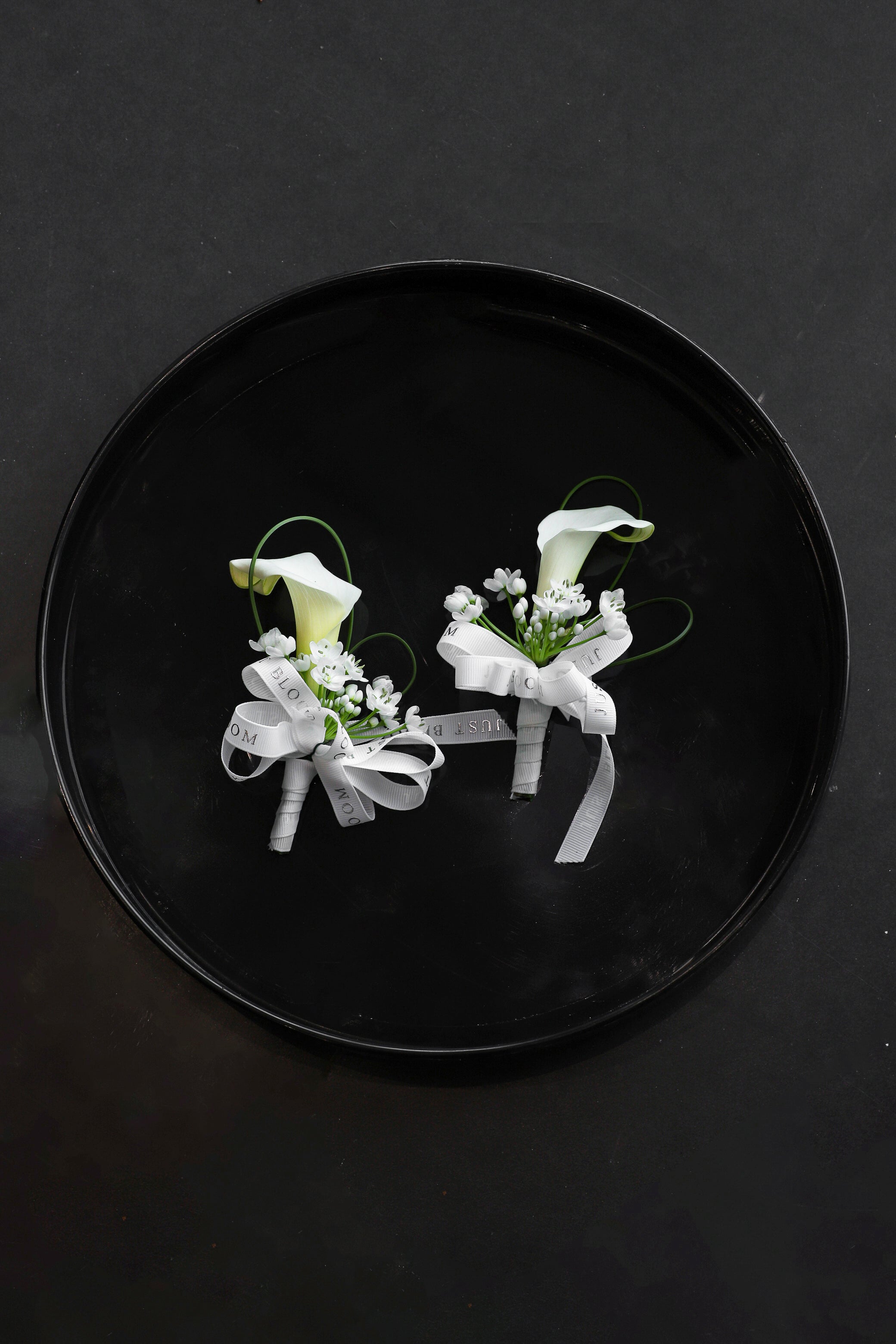 Two Boutonnieres arranged on a black tray featuring white calla lilies, allium cowanii and spiral steel grass tied with Just Bloom white ribbon — Just Bloom Hong Kong