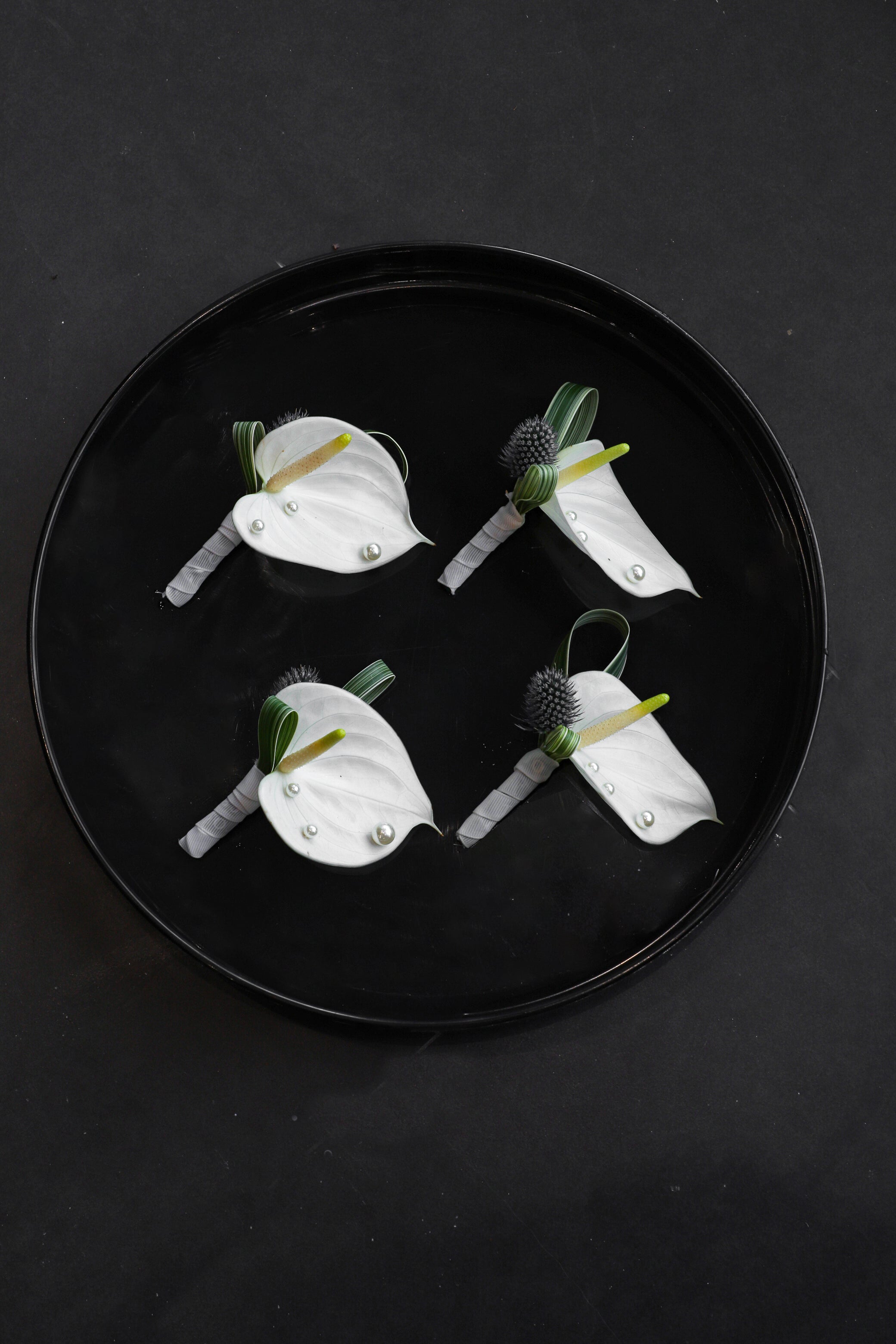 Four Boutonnieres arranged on a black tray featuring white anthuriums, blue globe thistles, foliage and pearl accents tied with Just Bloom grey ribbon — Just Bloom Hong Kong