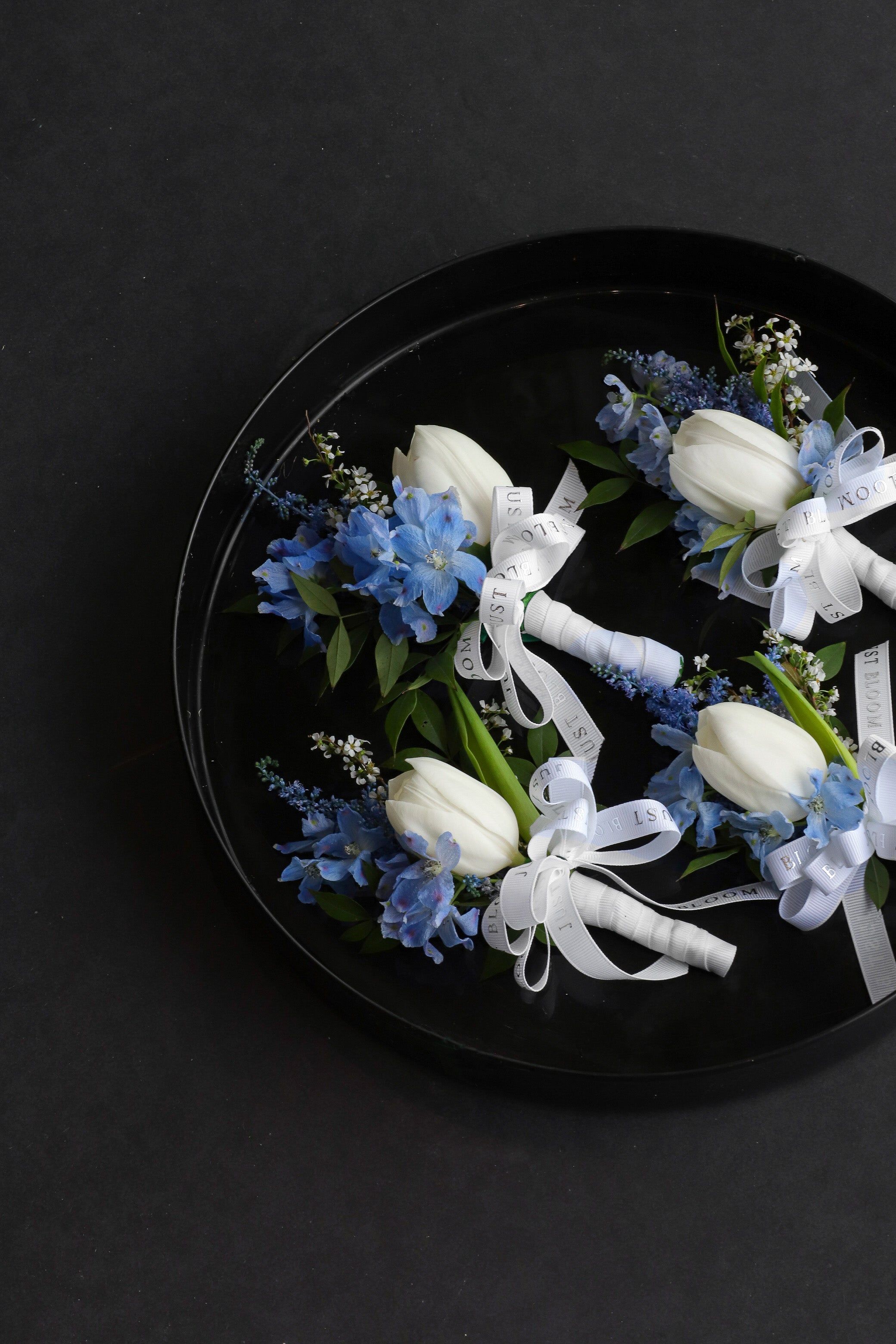 Four Blue Boutonnieres arranged on a black tray featuring white tulips, dusty blue delphiniums and astilbes tied with Just Bloom white ribbon — Just Bloom Hong Kong