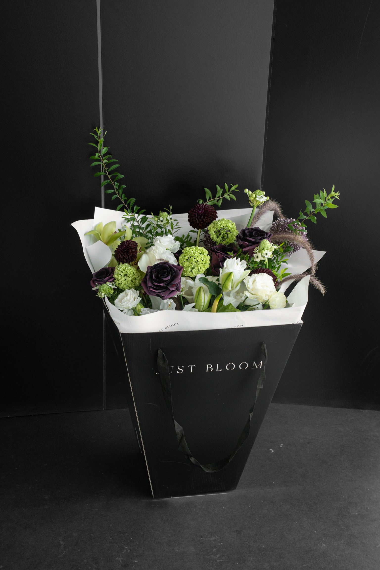 Just Bloom signature black gift bag carrying a large dark-toned fresh flower bouquet with deep plum and lime green blooms against a dark background — Just Bloom Hong Kong