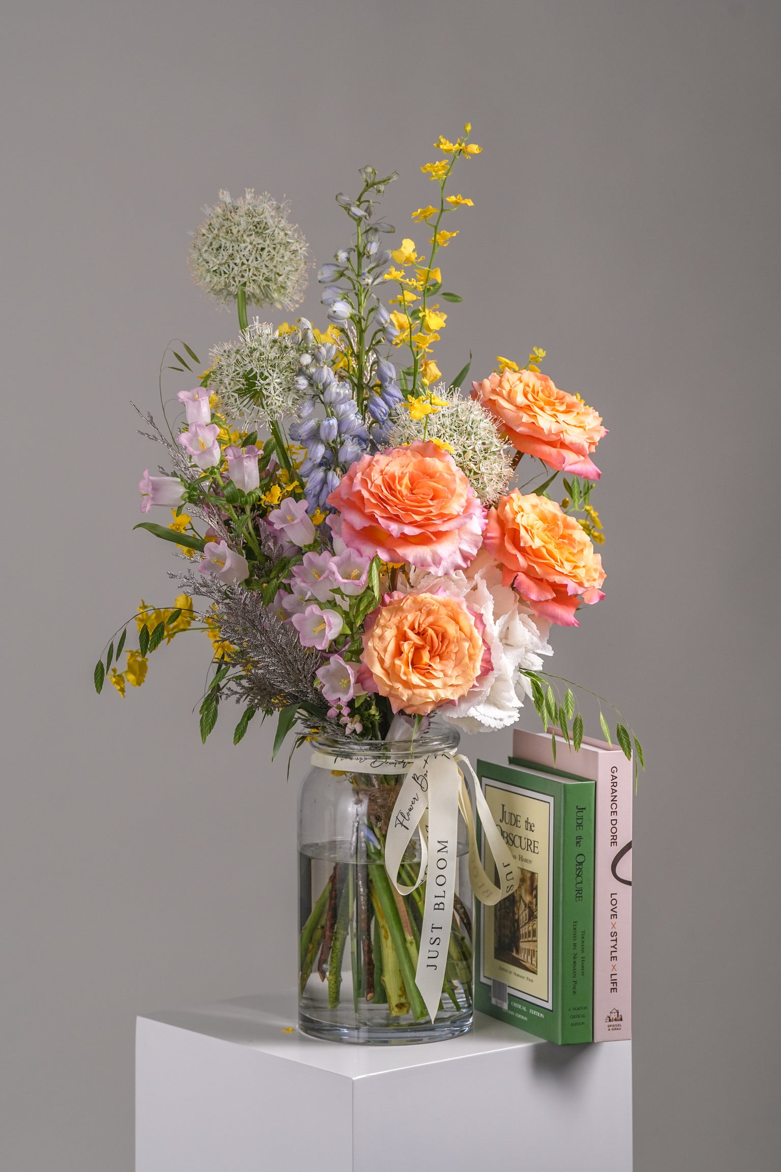 Colorful flower arrangement in a glass vase featuring orange Ecuadorian roses, Canterbury bells, giant alliums, and spring pastels beside books.