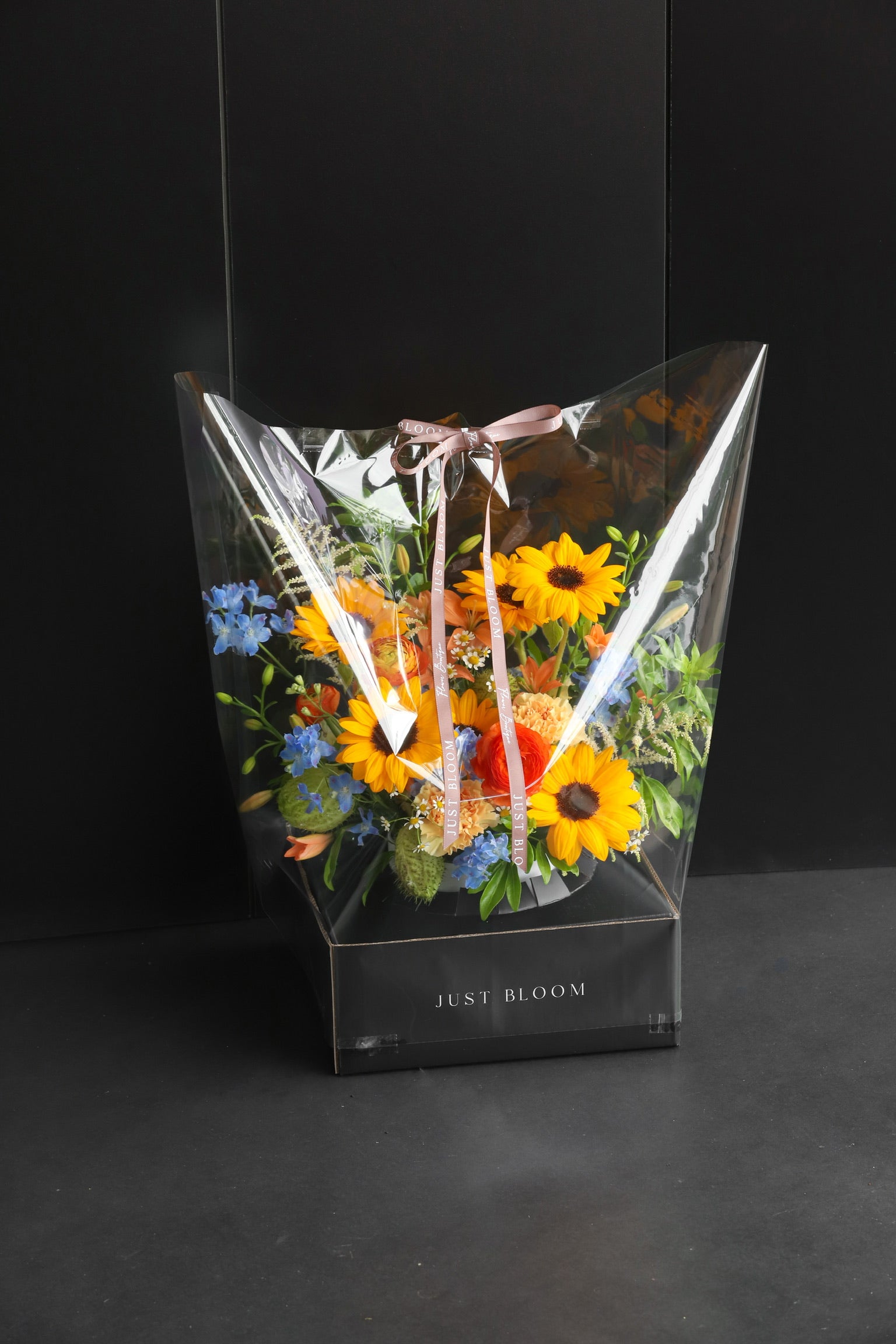 Just Bloom flower box arrangement in clear gift packaging with ribbon, showing warm-toned sunflowers and mixed blooms against a dark background — Just Bloom Hong Kong