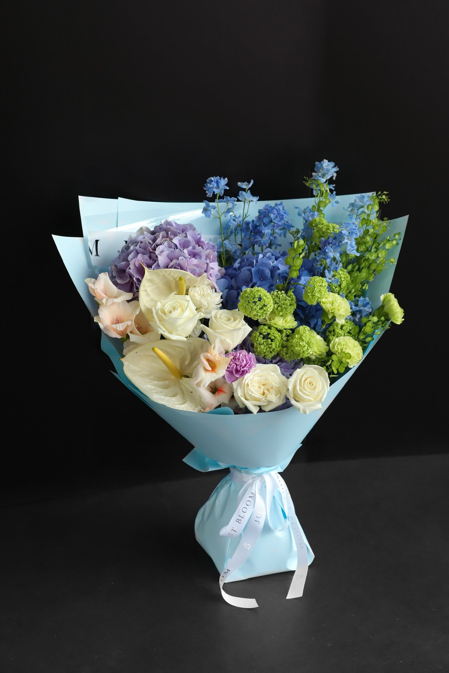 Large fresh flower bouquet in layered blue and purple tones wrapped in sky blue paper with white ribbon, photographed against a dark background — Just Bloom Hong Kong