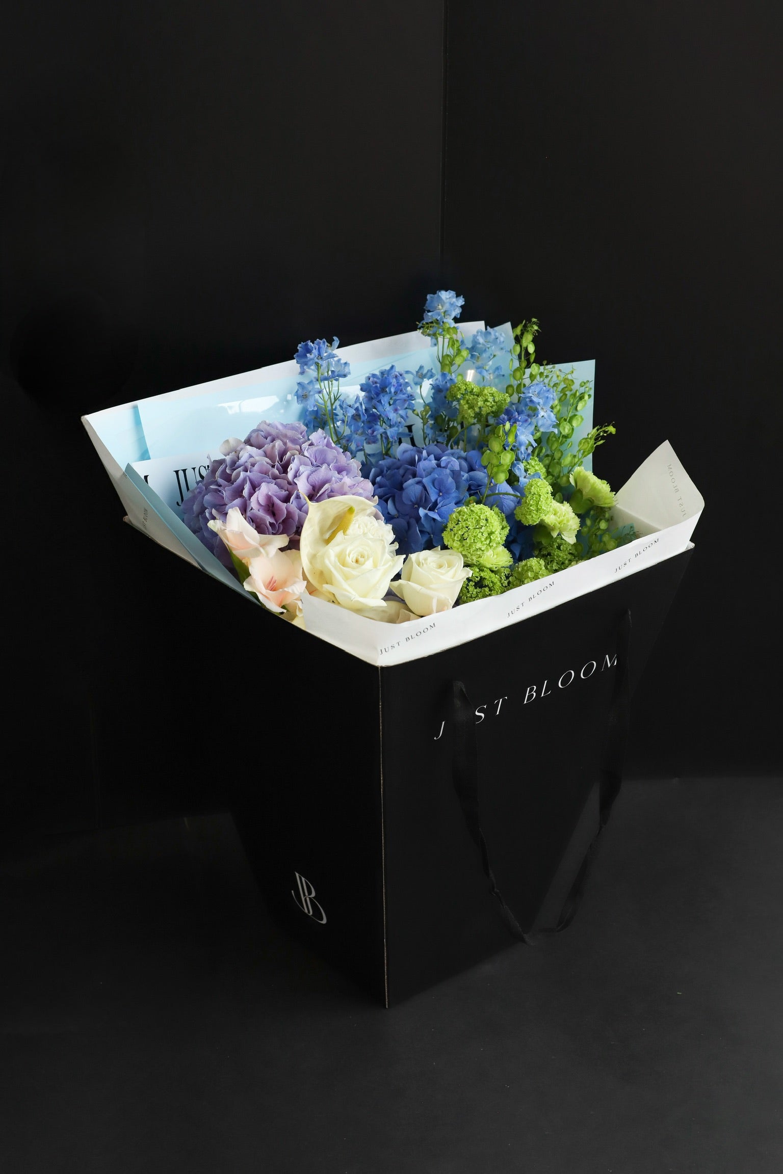 Just Bloom signature black gift bag carrying a large cool-toned fresh flower bouquet with blue and purple blooms against a dark background — Just Bloom Hong Kong
