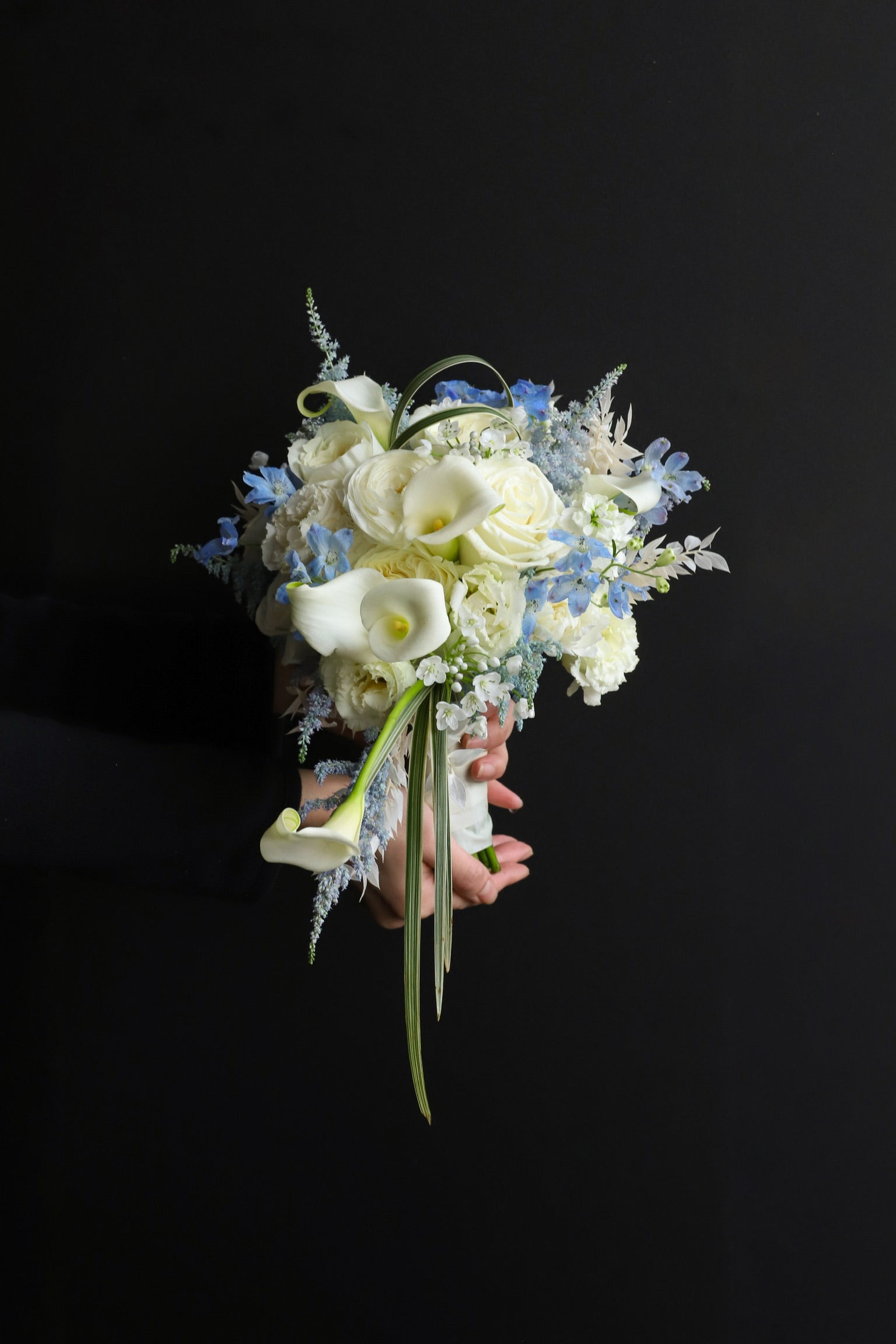 Close-up of a cascading bridal bouquet in white and blue tones with trailing calla lilies and leaf blades, held against a dark background — Just Bloom Hong Kong