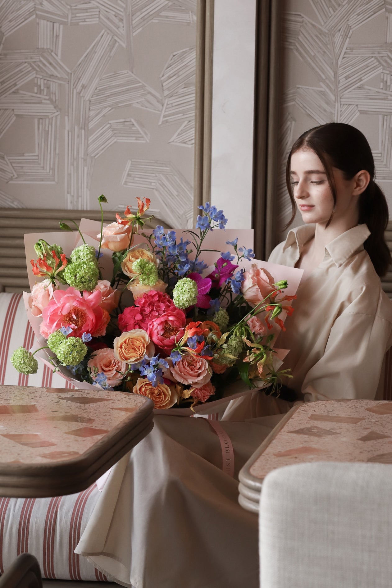 A model seated in a luxury restaurant setting holding Blooming with Love bouquet by Just Bloom Hong Kong, featuring an abundance of colourful blooms in warm peach, coral, pink and blue tones
