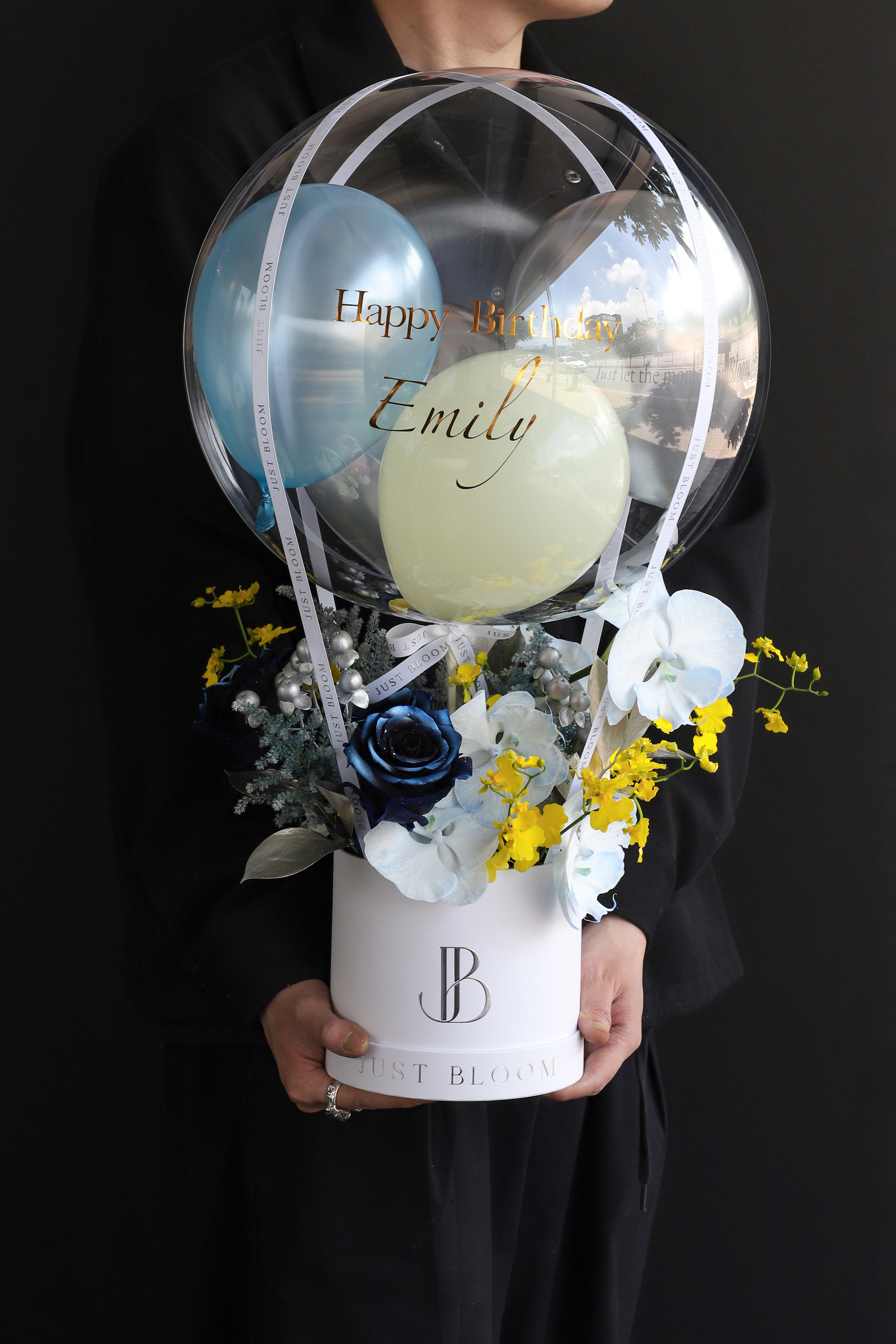 Model holding a fresh flower box with navy tinted Roses, white Orchids, and yellow Dancing Orchids in a white Just Bloom round box against a dark background — Just Bloom Hong Kong