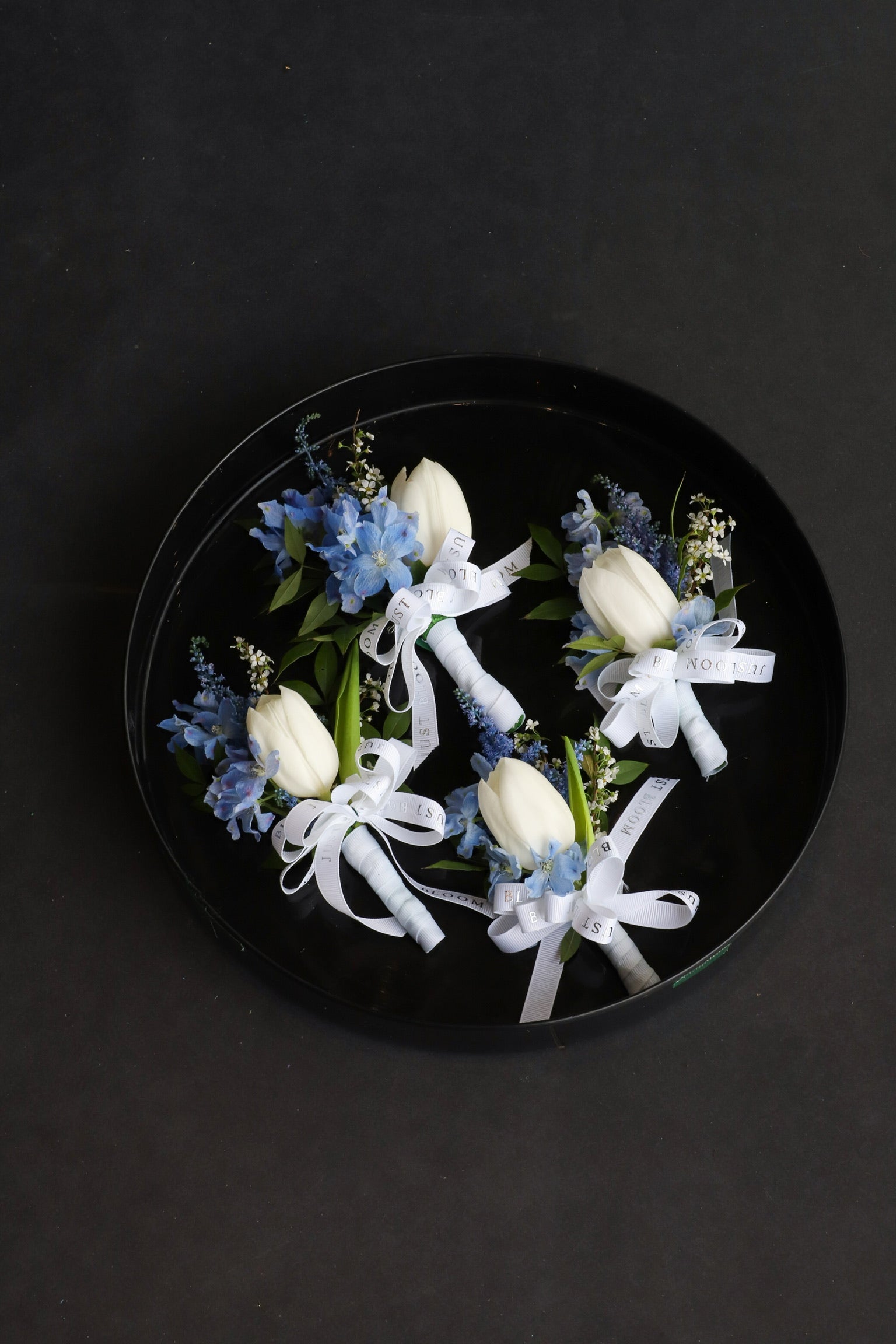 Three Boutonnieres displayed on a black tray by Just Bloom Hong Kong, showcasing white tulips with soft blue delphiniums, astilbes and white seasonal fillers