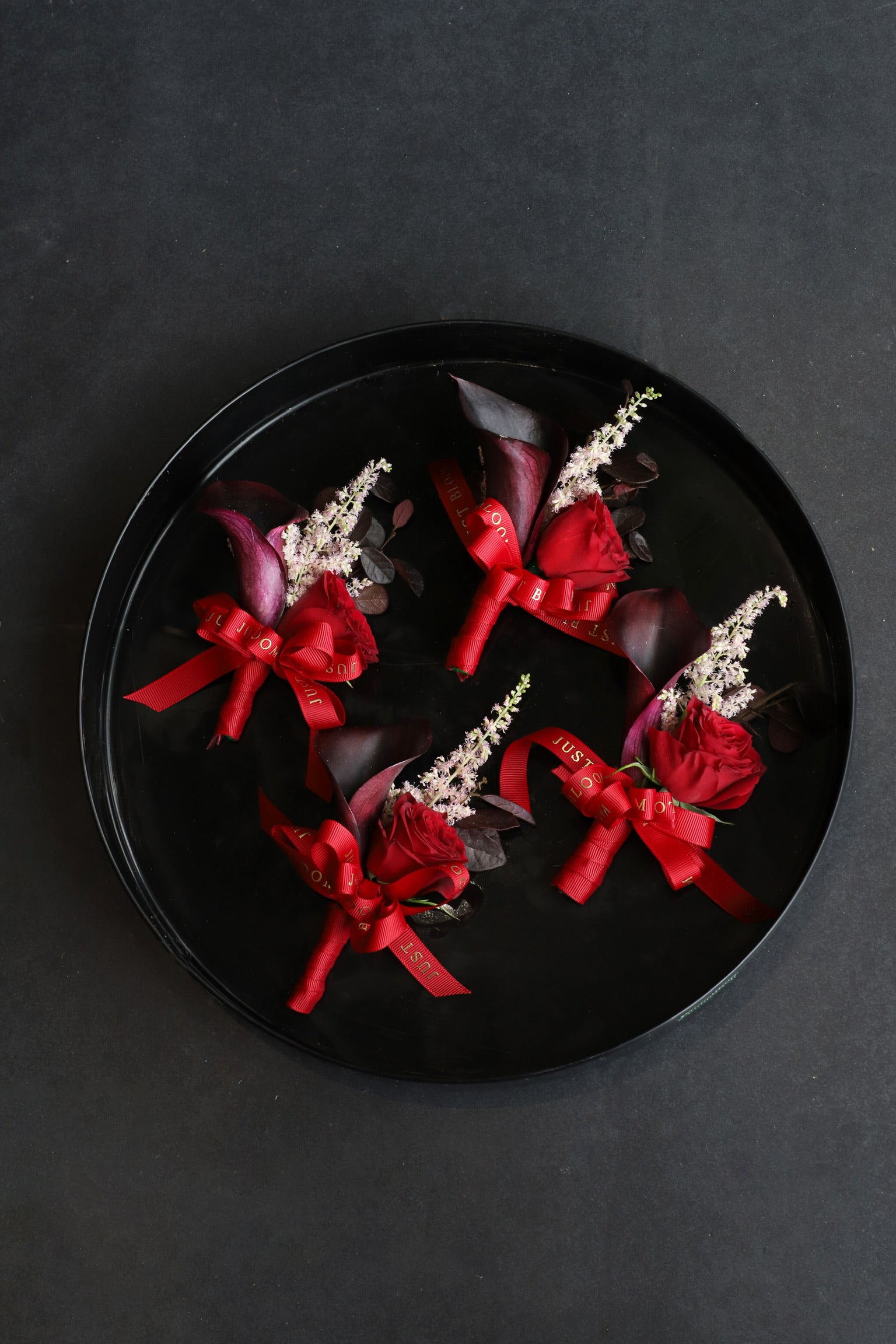 Four Bold Promise wedding corsages displayed on a black tray by Just Bloom Hong Kong, showcasing deep red spray roses, dark calla lilies, astilbes and Just Bloom red ribbon