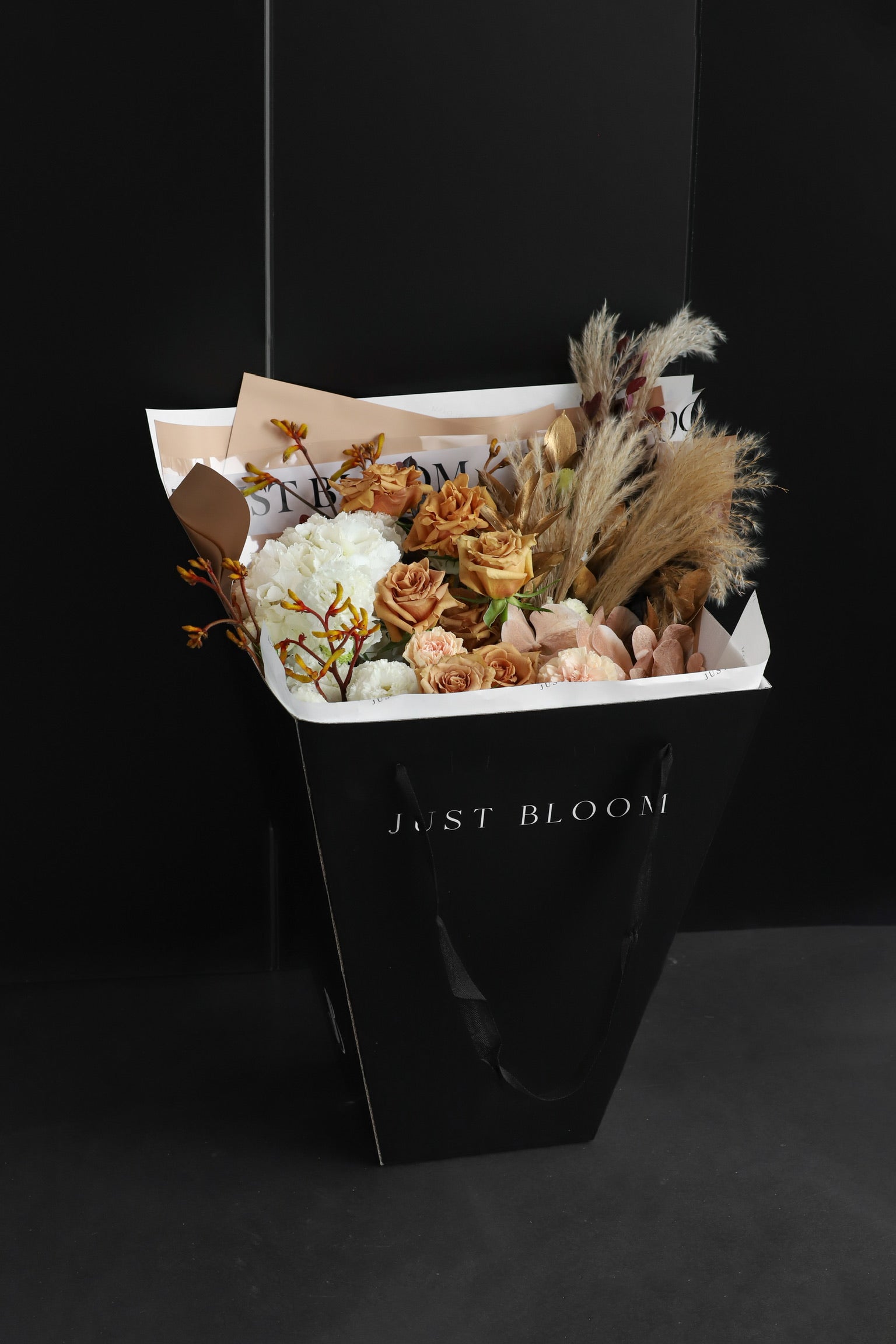 Just Bloom signature black gift bag carrying a large warm earth-toned bouquet with roses, white blooms, and dried pampas elements against a dark background — Just Bloom Hong Kong