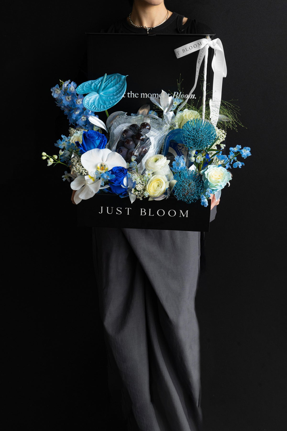 Model holding a large all-blue luxury floral hamper with premium fruits in a black Just Bloom box against a dark background — Just Bloom Hong Kong