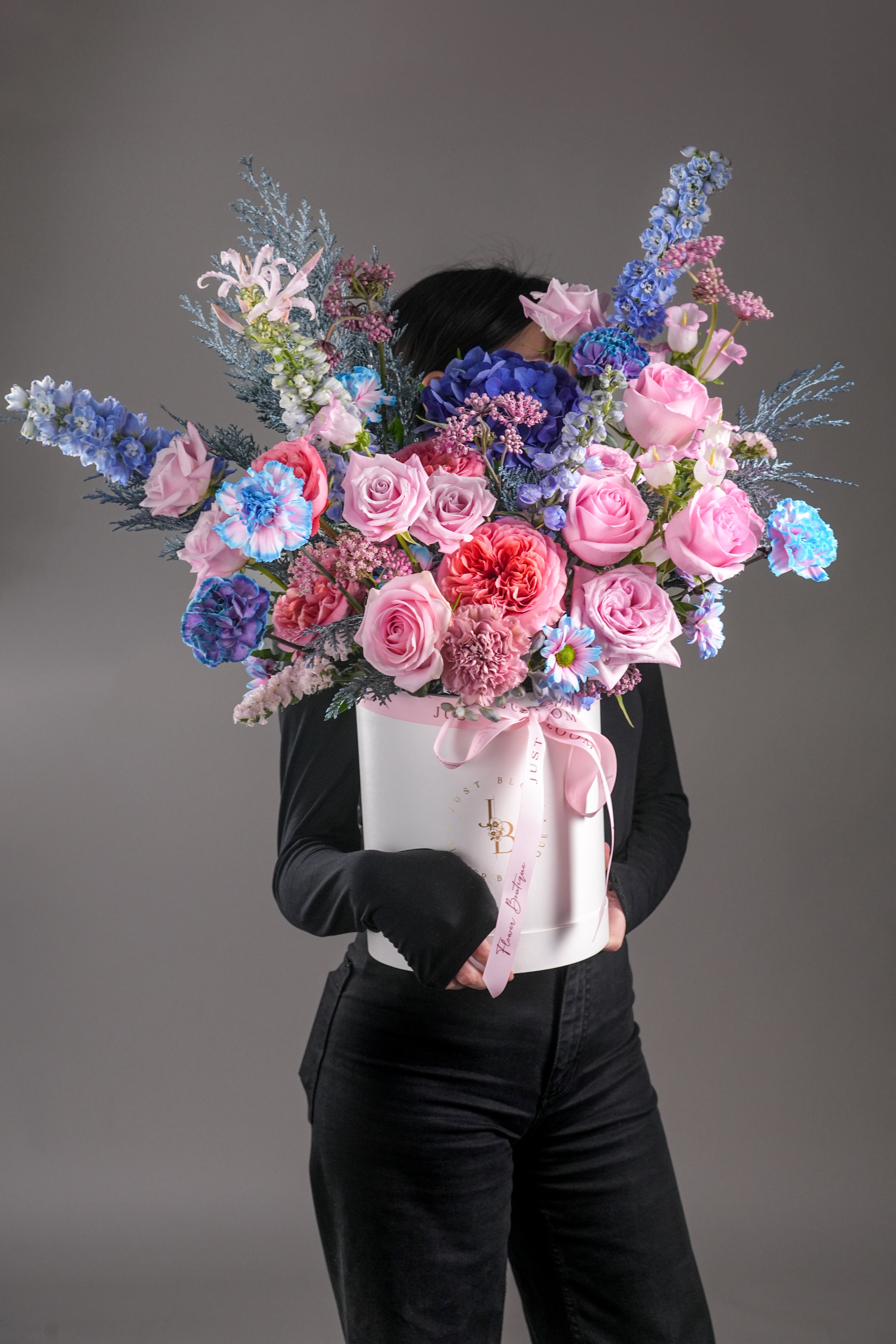 Model holding arrangement: Large pink and blue bouquet hatbox held by person in black – Ella