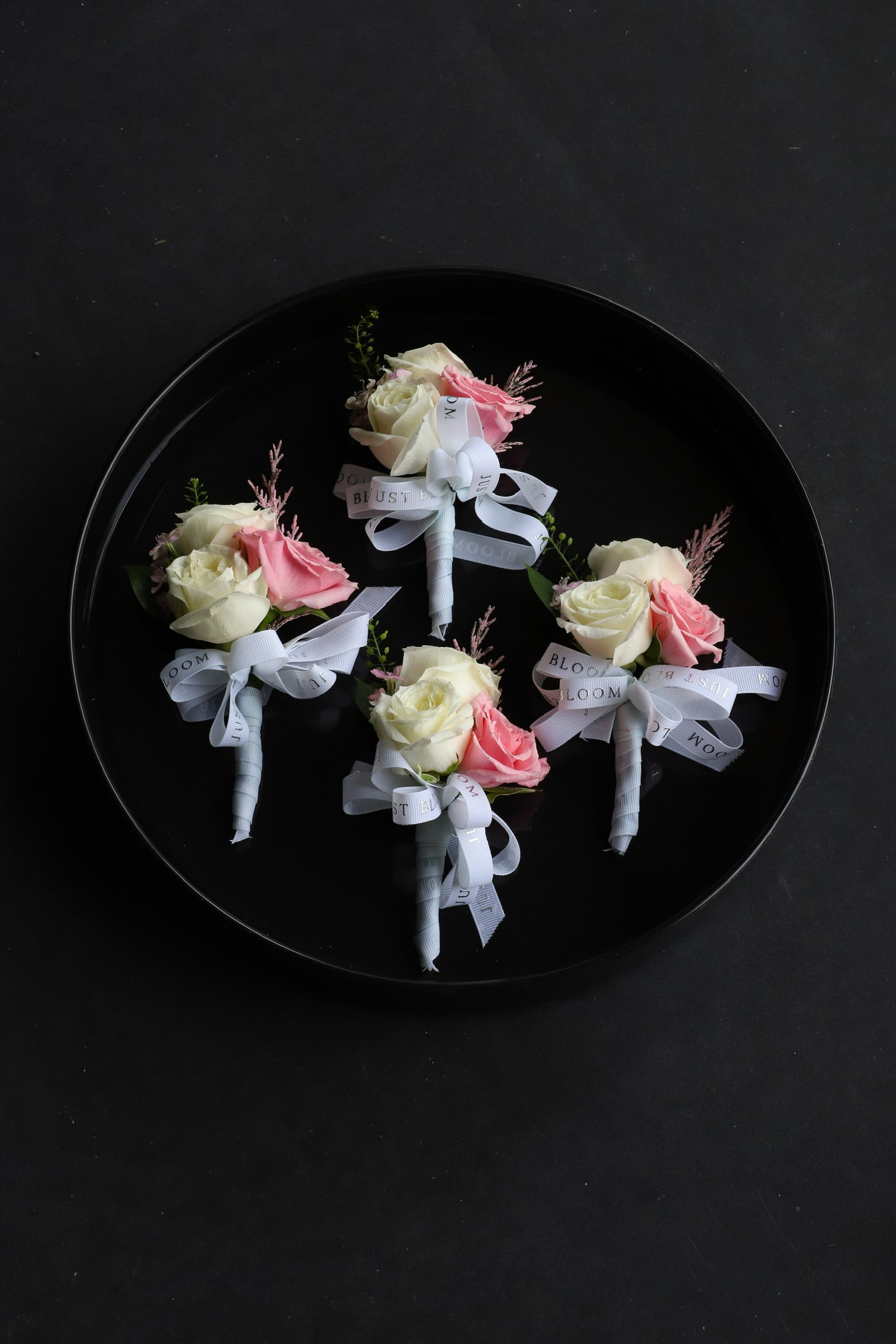 Four Boutonnieres arranged on a black tray featuring white and pink Ecuadorian roses, frosted explosion and astilbes tied with Just Bloom white ribbon — Just Bloom Hong Kong