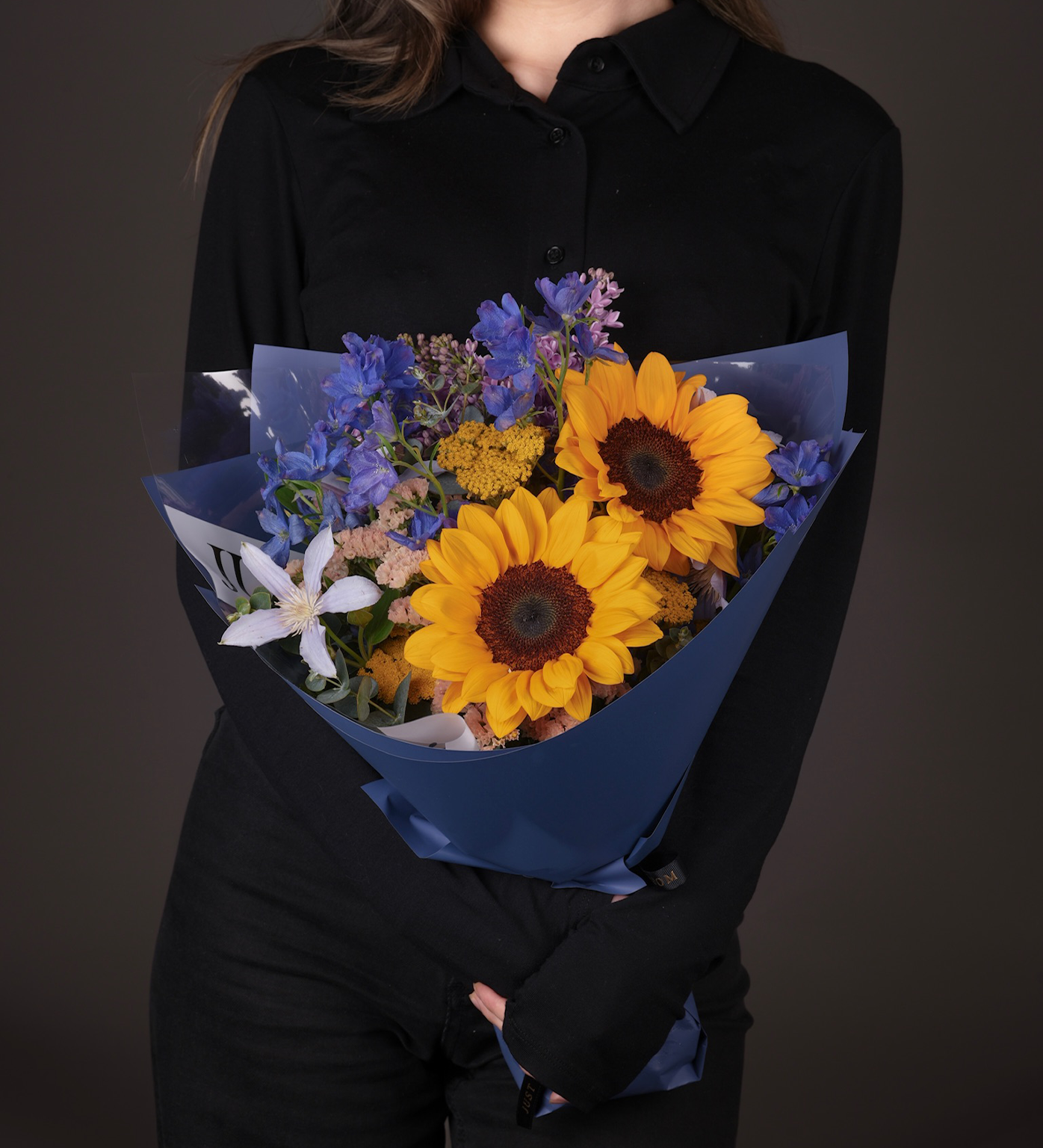 Kelsey sunflower and clematis mixed bouquet with blue and purple flowers in navy wrapping held by woman