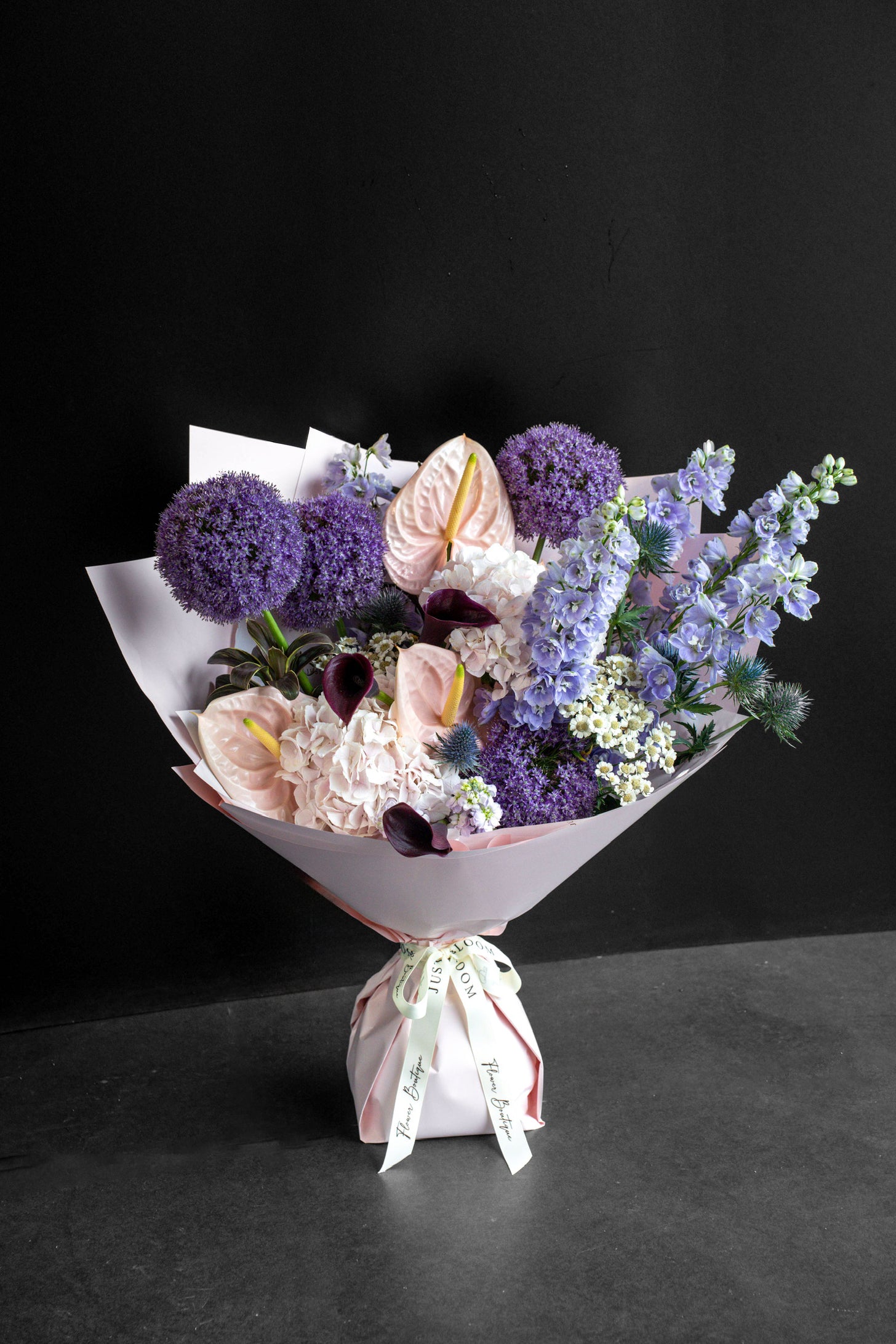 Lavender Muse bouquet of purple and plum blooms with blush accents wrapped in pink paper against a black backdrop — Just Bloom Hong Kong