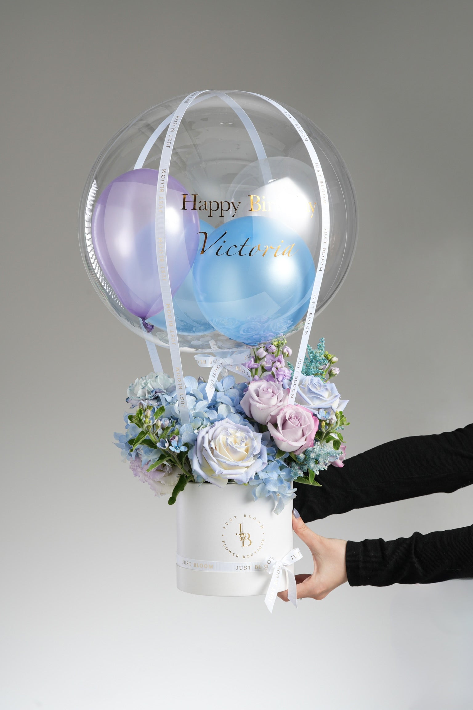 Hands holding Lydia flower box showing blue and purple roses with delphinium arrangement and birthday balloon