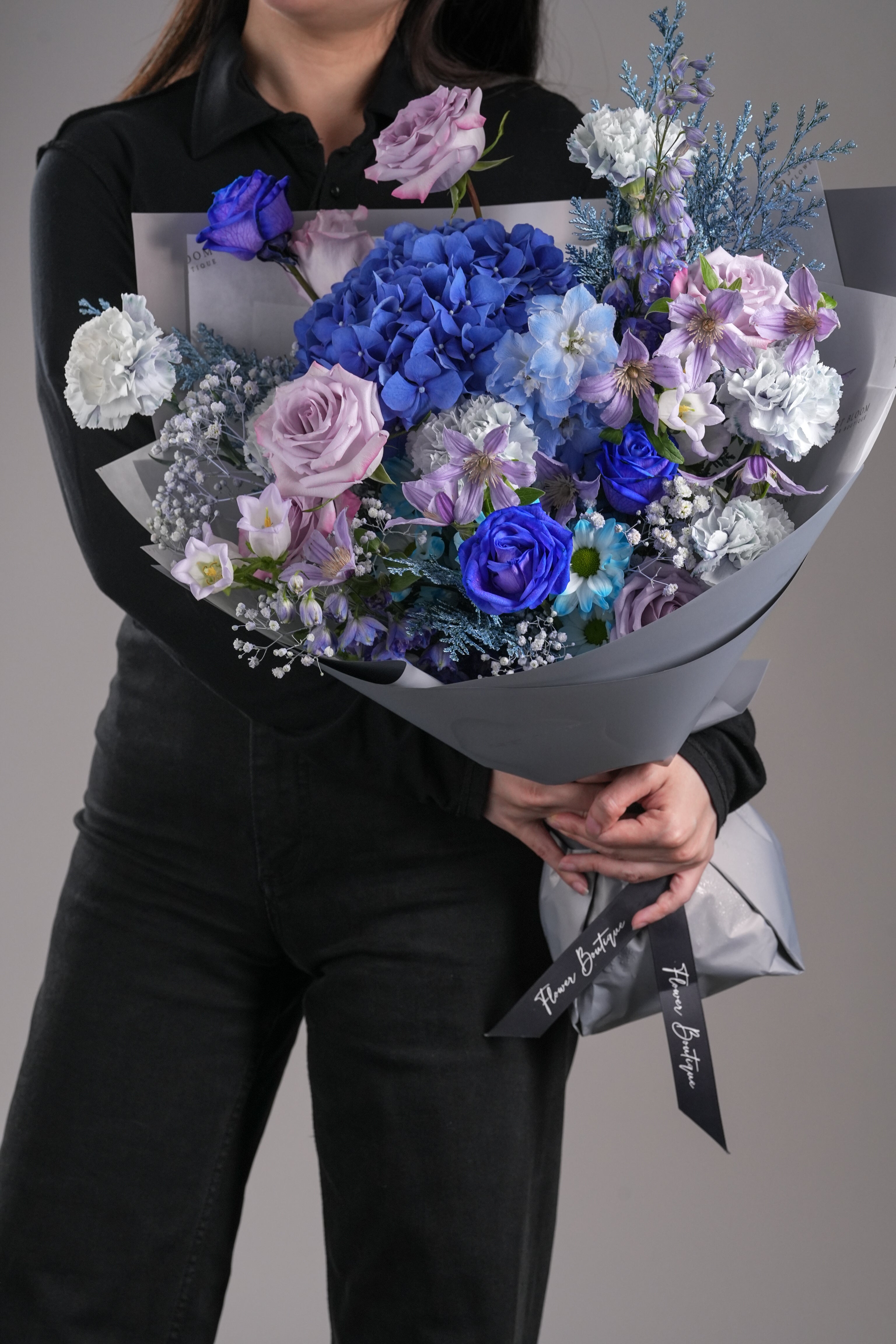 Maeve bouquet with blue hydrangeas, purple roses, white carnations, and flower fillers wrapped in grey-blue paper