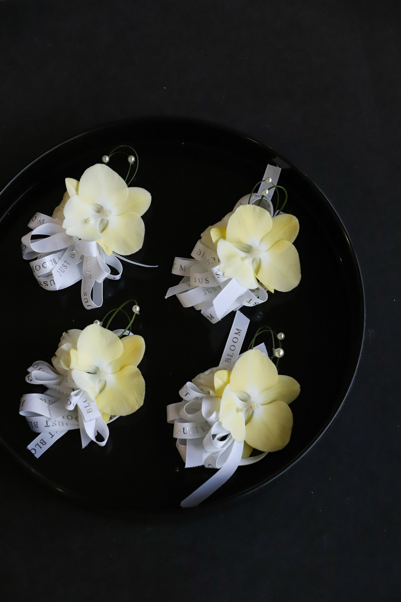 Close-up of four Meant to Be wedding corsages on a black tray by Just Bloom Hong Kong, showcasing cream phalaenopsis orchids with spiral steel grass, pearl accents and Just Bloom white ribbon