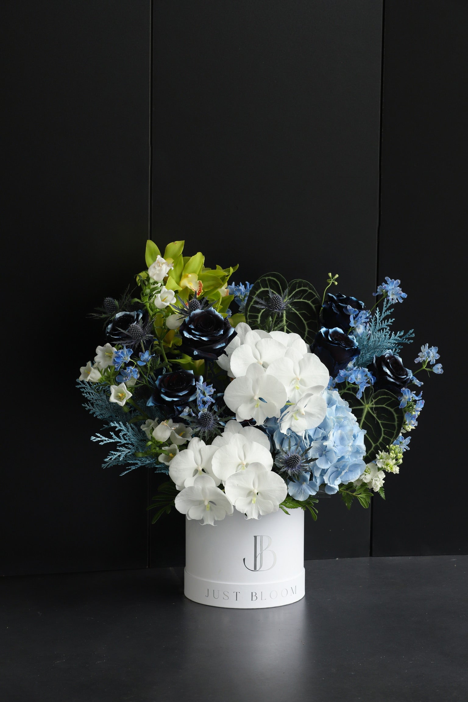 Dramatic deep navy and white flower box arrangement with Cymbidium Orchids, dyed Roses, and mixed seasonal blooms in a white Just Bloom hat box against a dark background — Just Bloom Hong Kong