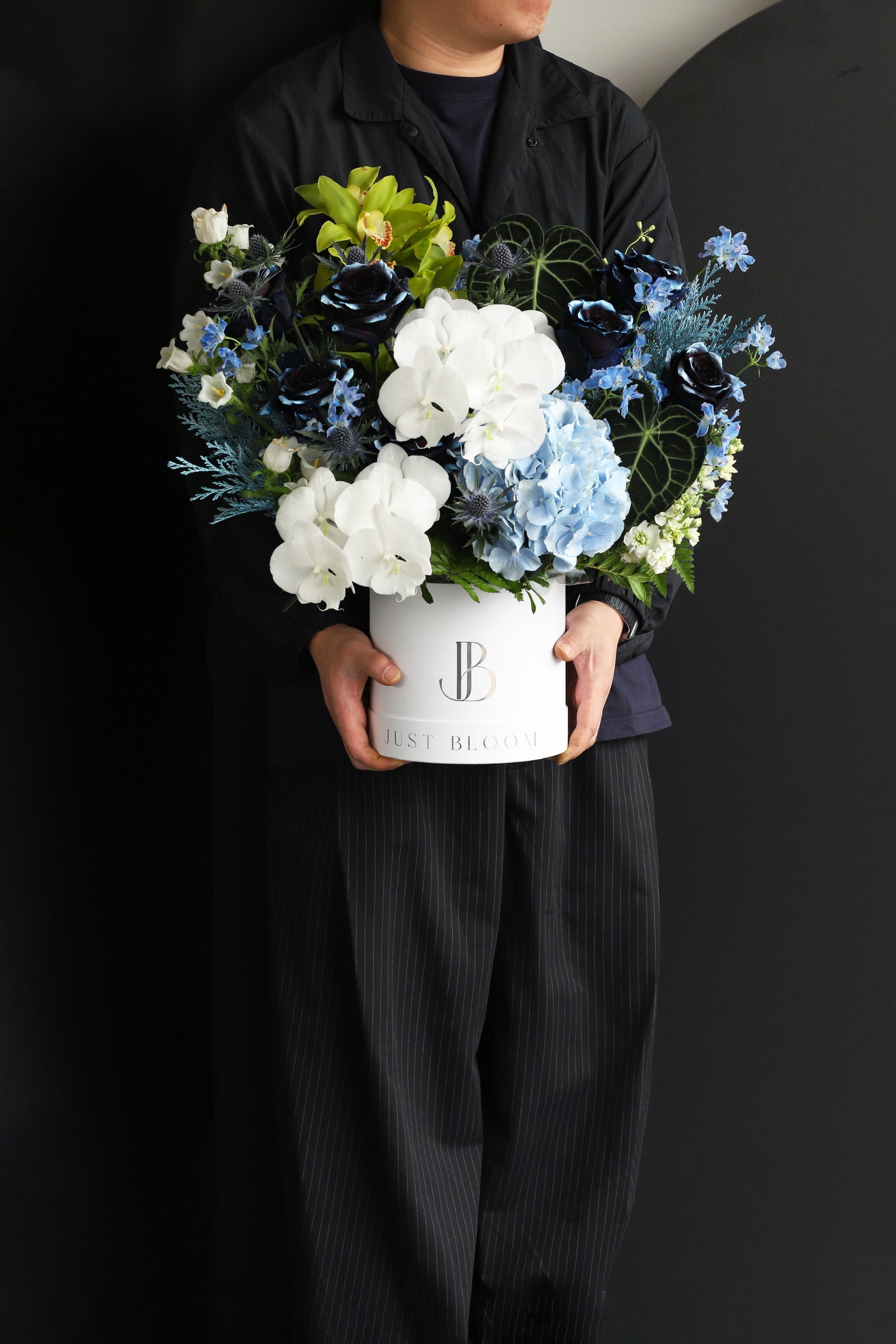 Model holding a large dramatic flower box arrangement in deep navy, white, and green tones with Cymbidium Orchids and mixed seasonal blooms in a white Just Bloom hat box — Just Bloom Hong Kong