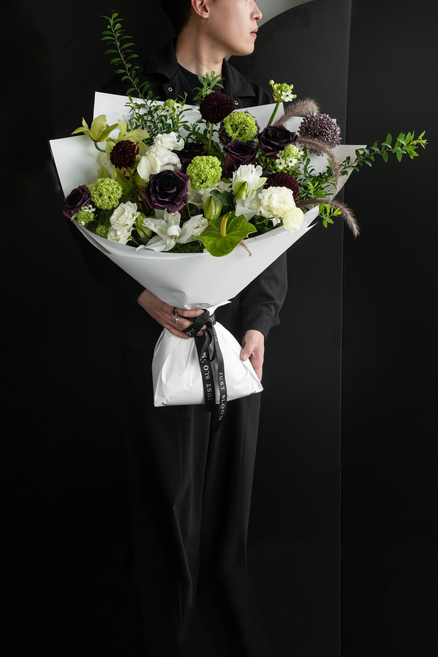 Model holding an oversized dark-toned fresh flower bouquet in deep plum and lime green tones wrapped in white Just Bloom paper against a slatted wall — Just Bloom Hong Kong