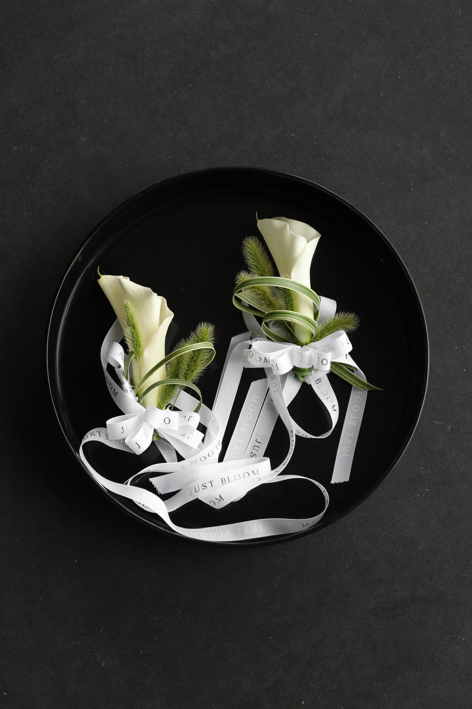 Two Pure Intention wrist corsages arranged on a black tray featuring white calla lilies, spider plants and foxtail grass with Just Bloom white ribbon — Just Bloom Hong Kong