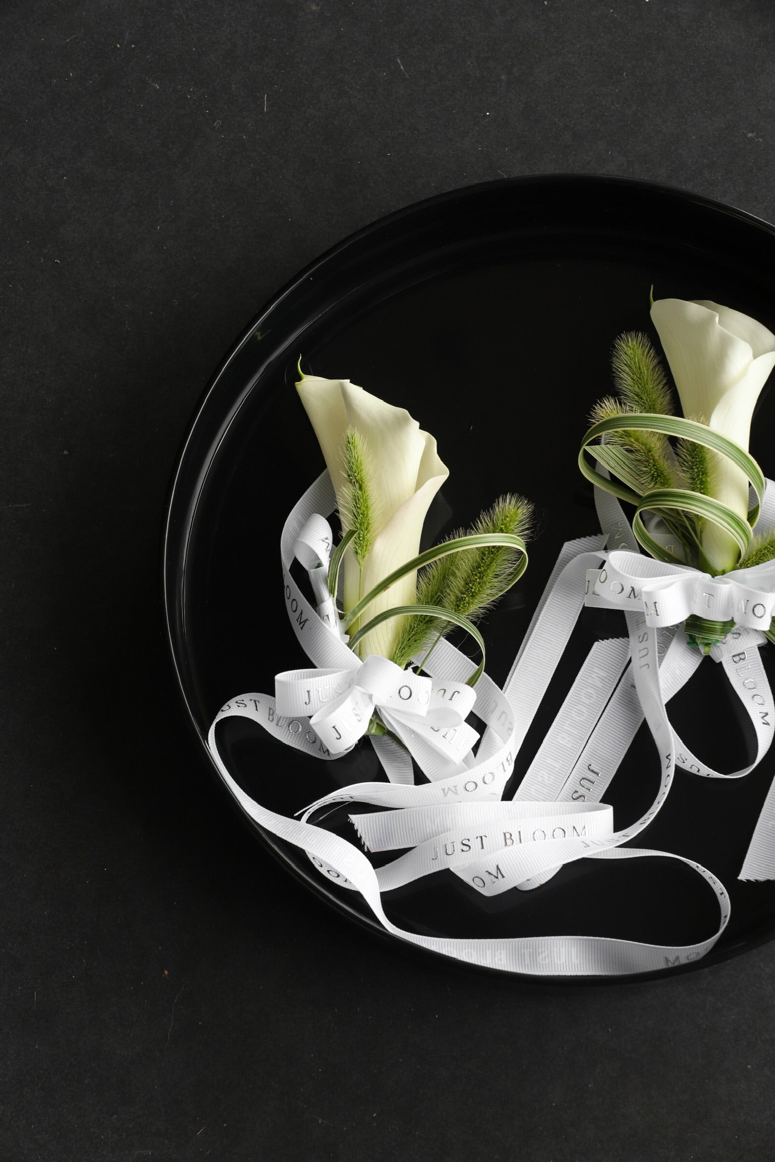 Close-up of two Pure Intention wedding wrist corsages on a black tray by Just Bloom Hong Kong, showcasing white calla lilies wrapped with spider plants, foxtail grass and Just Bloom white ribbon
