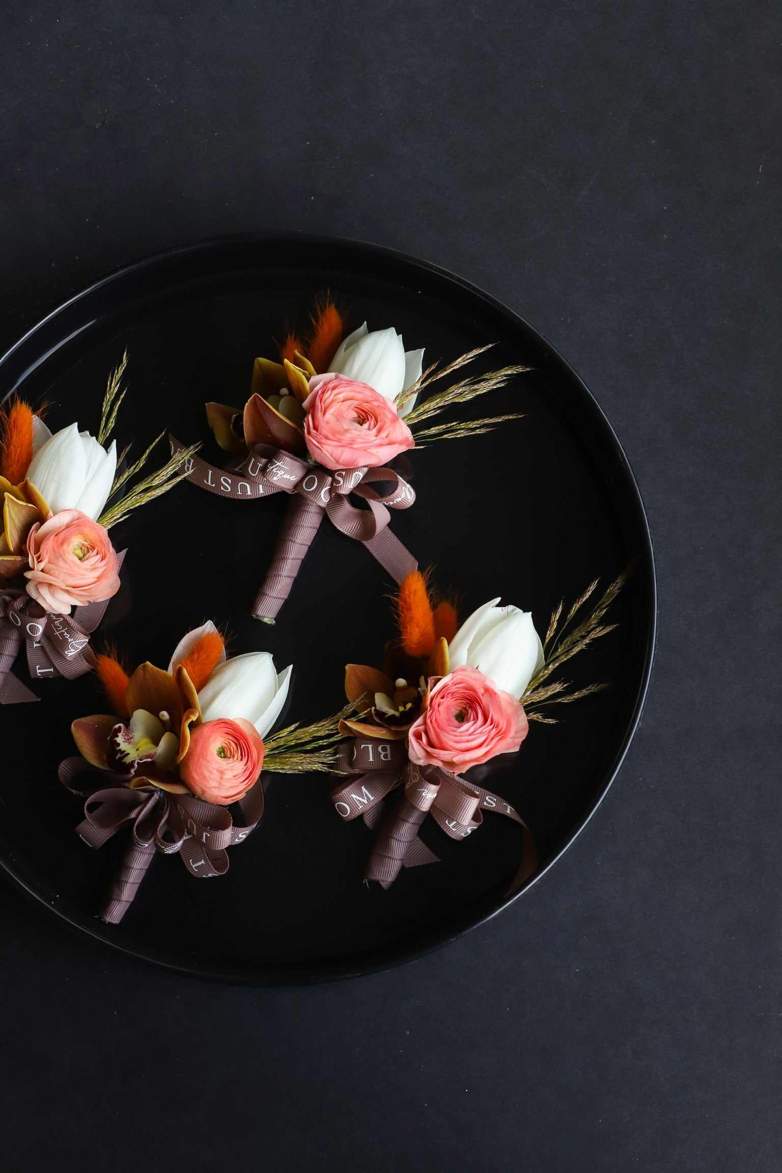 Close-up of four Boutonnieres on a black tray by Just Bloom Hong Kong, showcasing white tulips, coral ranunculus, cymbidium orchids, dried lagurus ovatus and Just Bloom dark ribbon