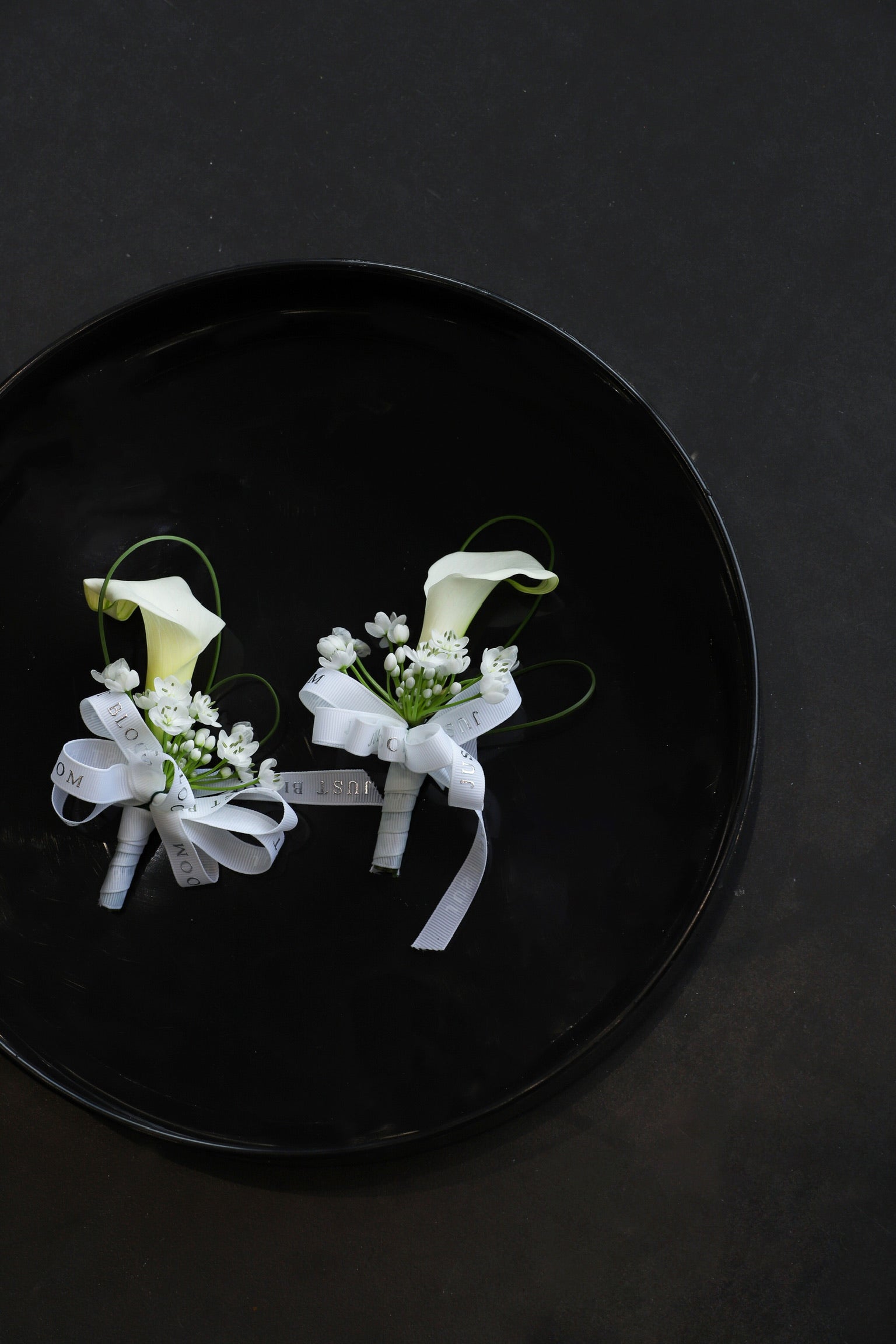 Close-up of two Simply White wedding corsages on a black tray by Just Bloom Hong Kong, showcasing white calla lilies, allium cowanii and delicate spiral steel grass with Just Bloom white ribbon