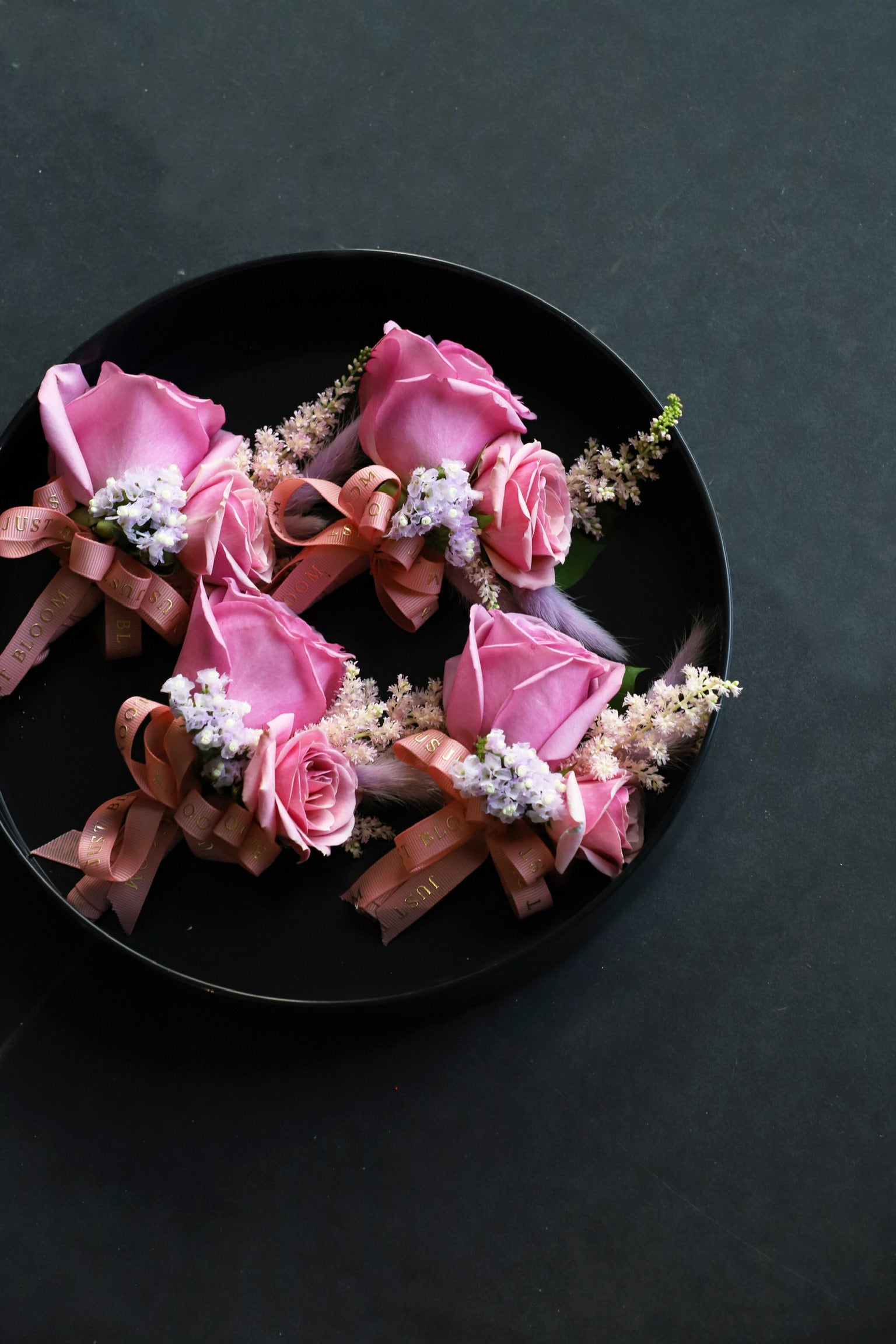 Close-up of four Boutonnieres on a black tray by Just Bloom Hong Kong, showcasing pink roses, pink astilbes, myosotis and feathery accents with Just Bloom rose gold ribbon
