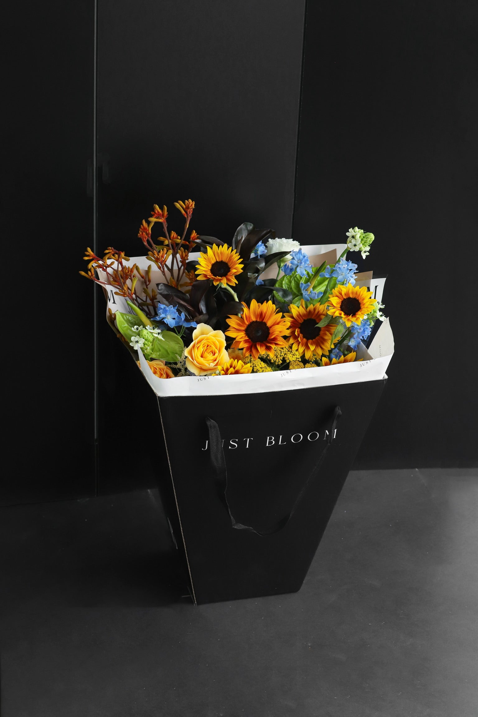Just Bloom signature black gift bag carrying a large fresh flower bouquet with sunflowers and warm-toned seasonal blooms — Just Bloom Hong Kong