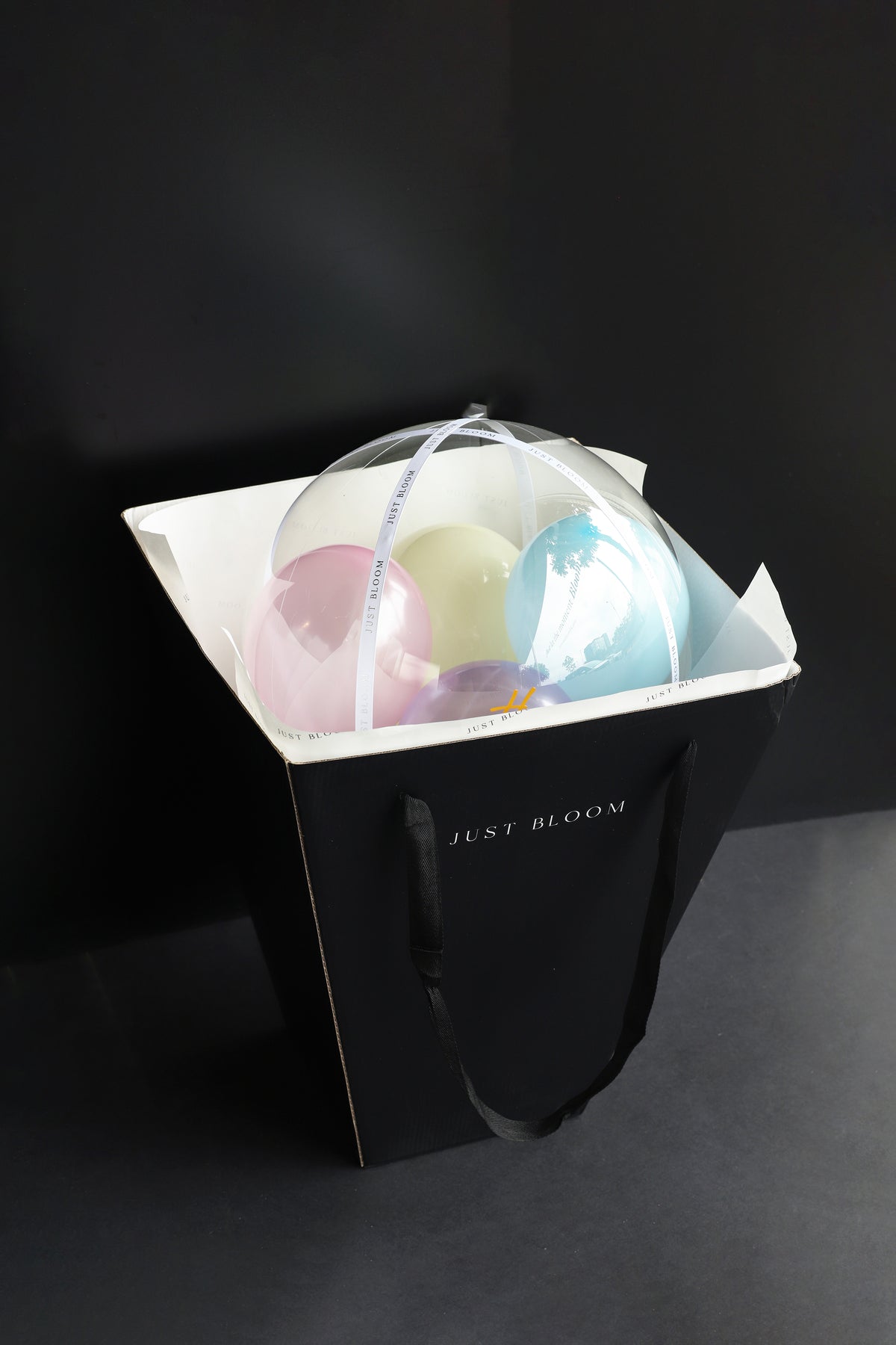 Just Bloom signature black gift bag carrying a Sugar Rush pastel flower box with multicoloured balloons visible through the crystal balloon against a dark background — Just Bloom Hong Kong