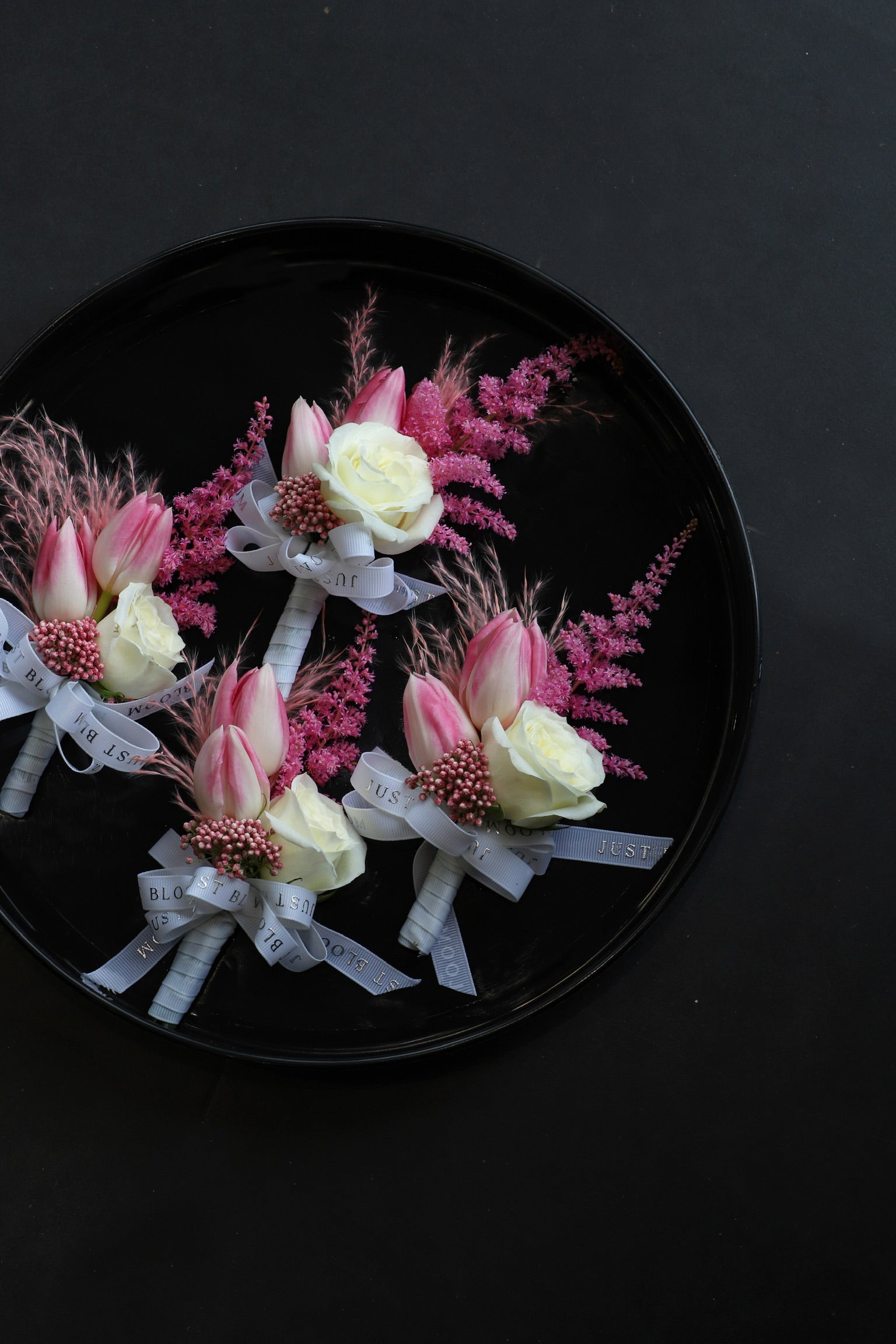 Close-up of four Sweet Vow Boutonnieres on a black tray by Just Bloom Hong Kong, showcasing pink tulips, white spray roses, deep pink astilbes and delicate rice flowers with Just Bloom grey ribbon